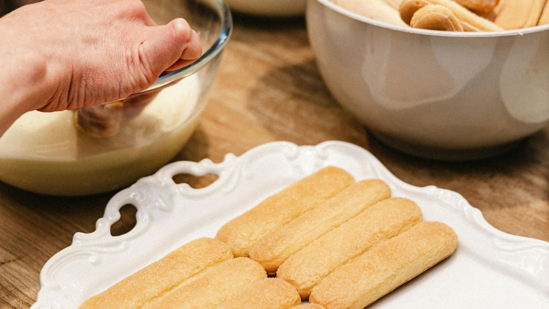 A person pours a liquid into a bowl, next to other ingredients and to a plate with sweets.
