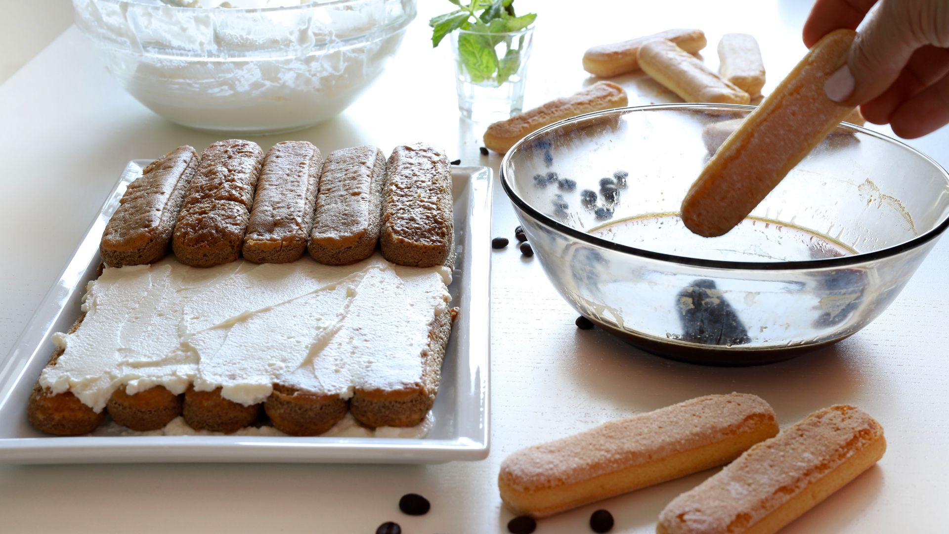 Picture of a table with ingredients and desserts for the preparation of tiramisu. In the foreground, a tray with tiramisu ready for tasting.