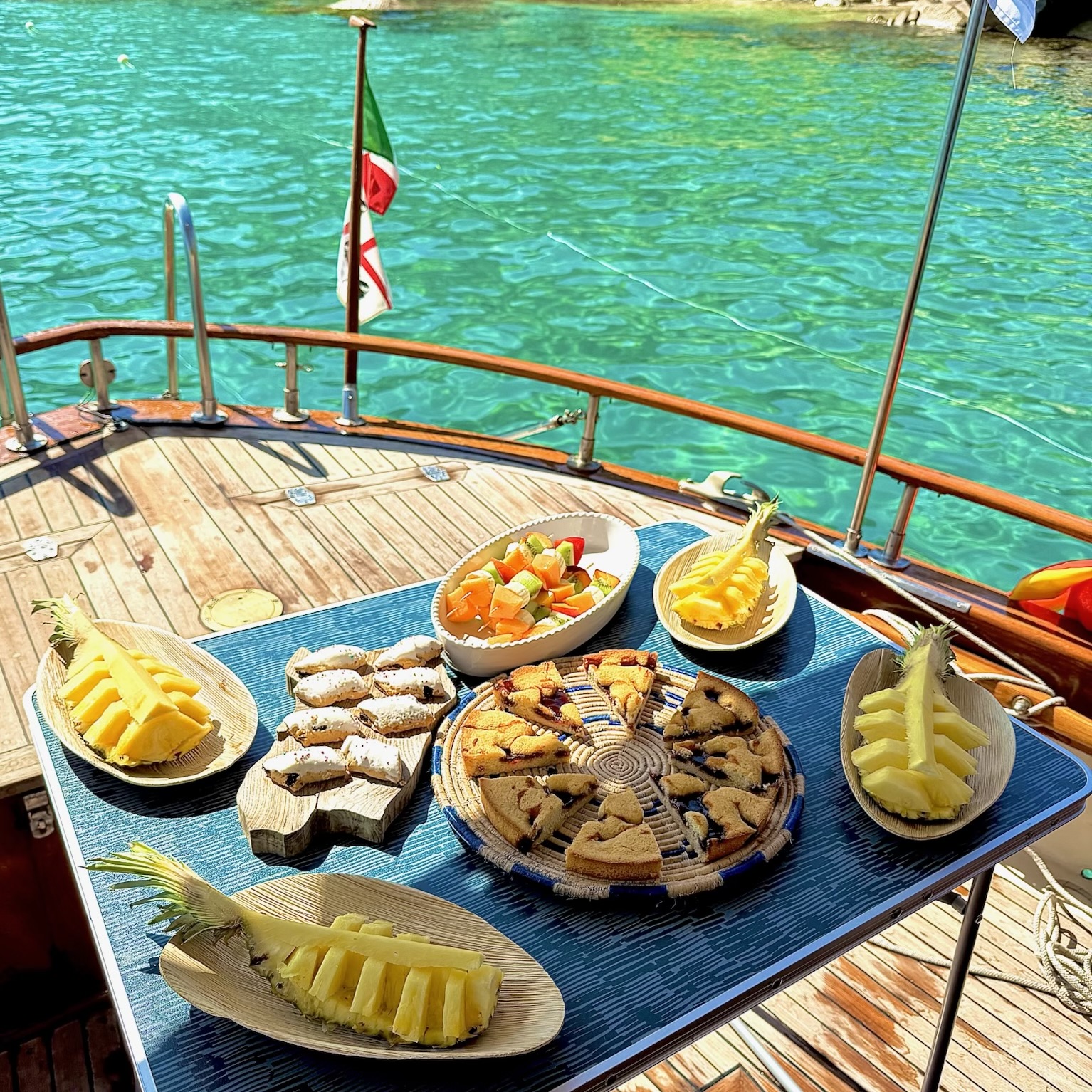 Ein Tisch mit Essen auf einem Segelboot mit blauem und grünem Meer im Hintergrund, einigen Inseln und einer Flagge.