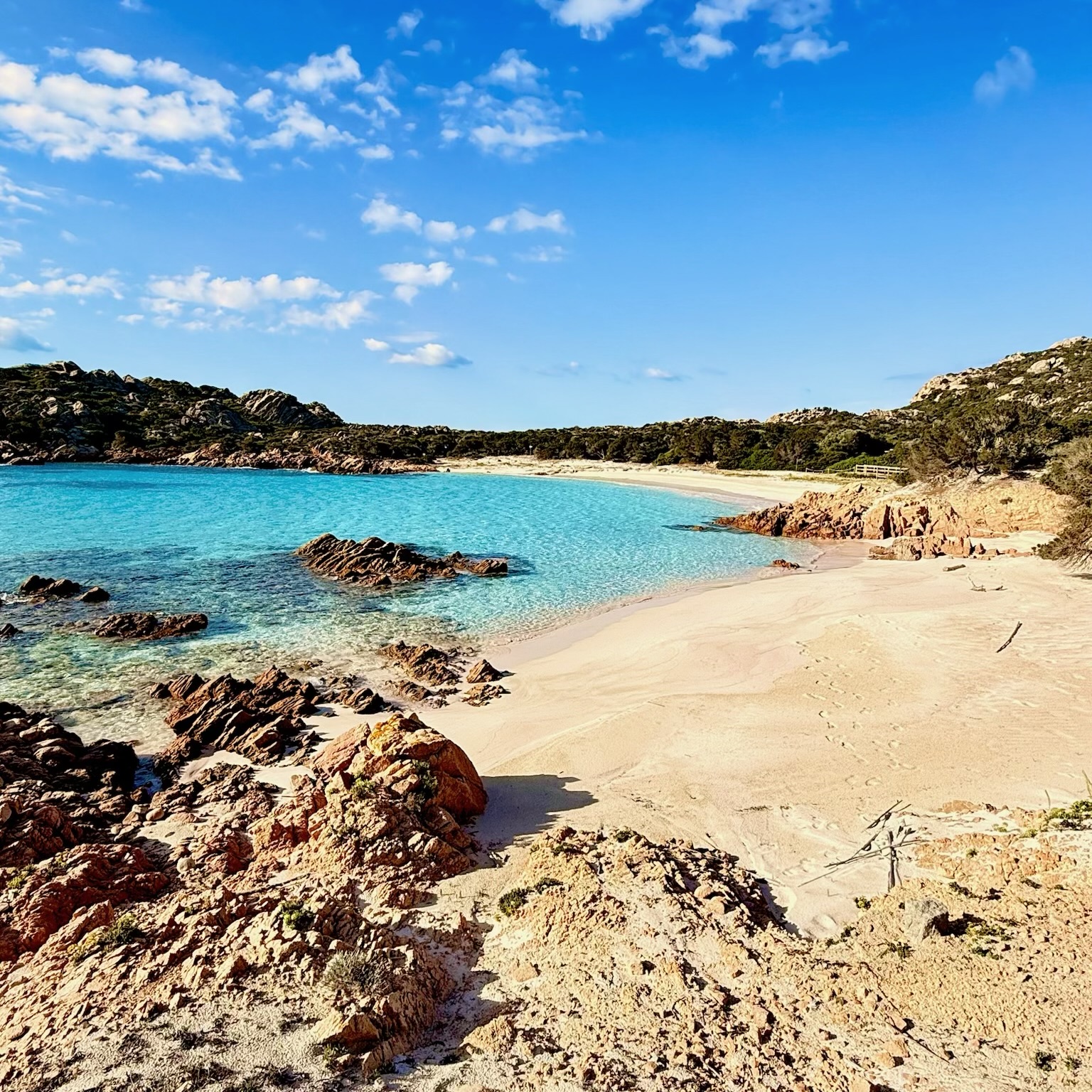 Strand mit kristallklarem Wasser und angrenzenden Felsen, unter blauem Himmel mit einigen Wolken.