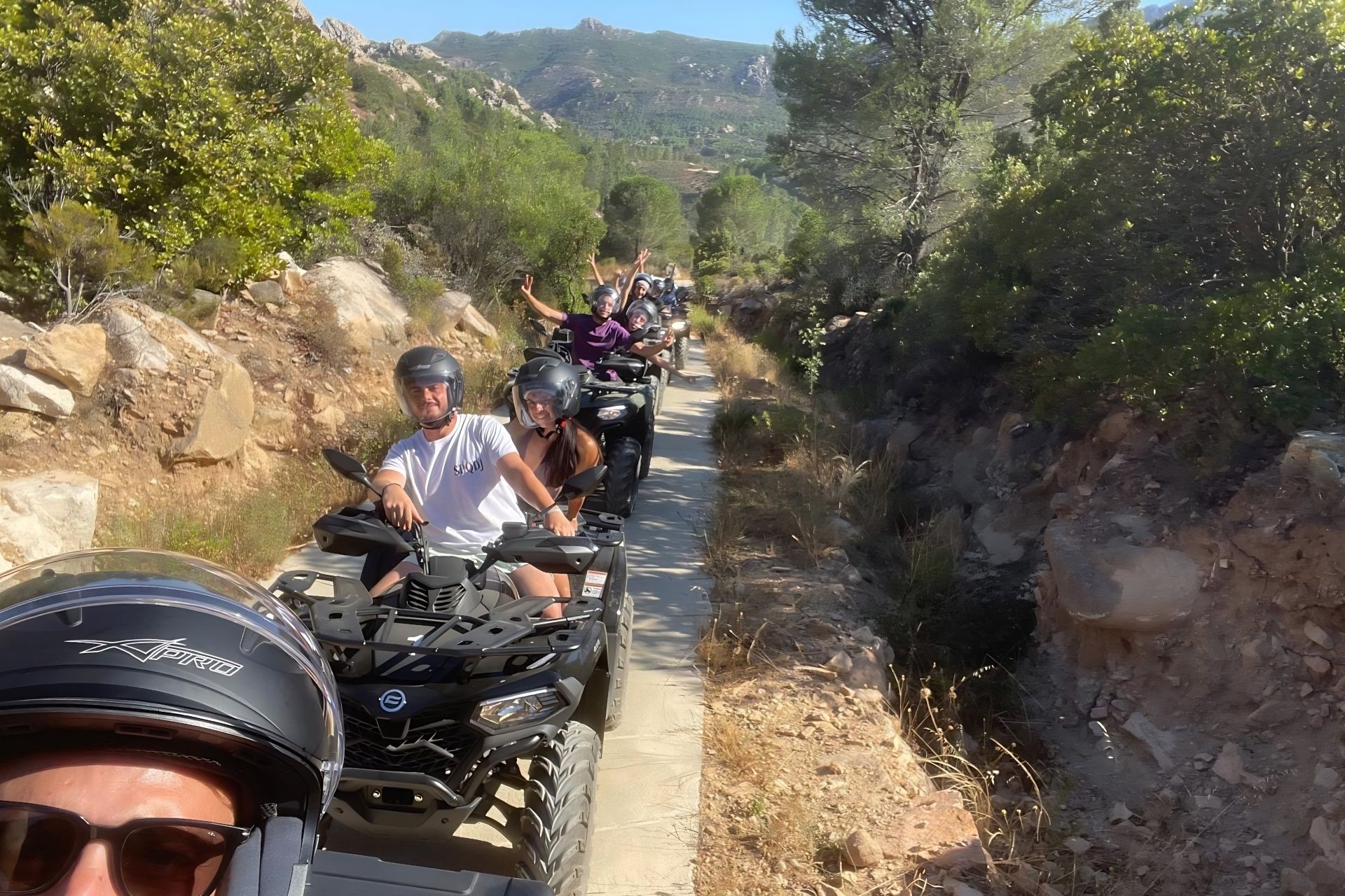 Groupe de personnes casquées conduisant un quad sur un chemin de terre, entouré de végétation et de rochers.
