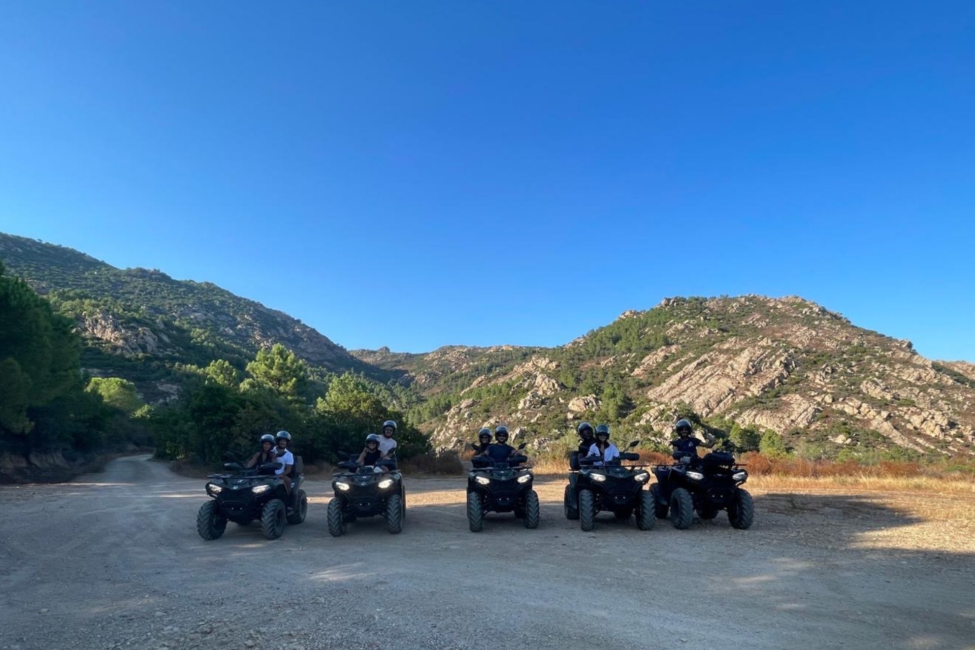 Groupe de personnes sur des quads dans un paysage montagneux, avec des montagnes et une végétation verte en arrière-plan. Tout le monde porte des casques et des gilets de sécurité.
