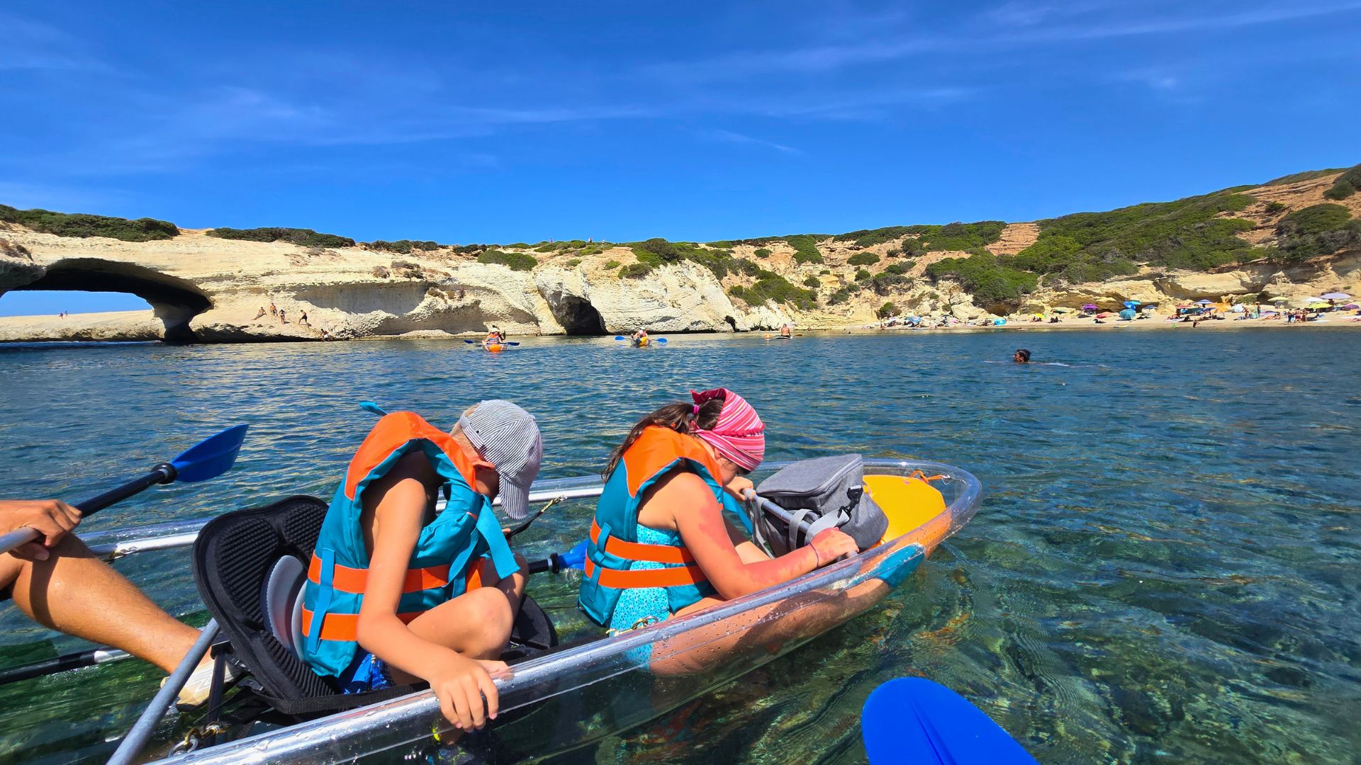 Due persone in un kayak trasparente vicino alla costa, con un arco naturale sullo sfondo