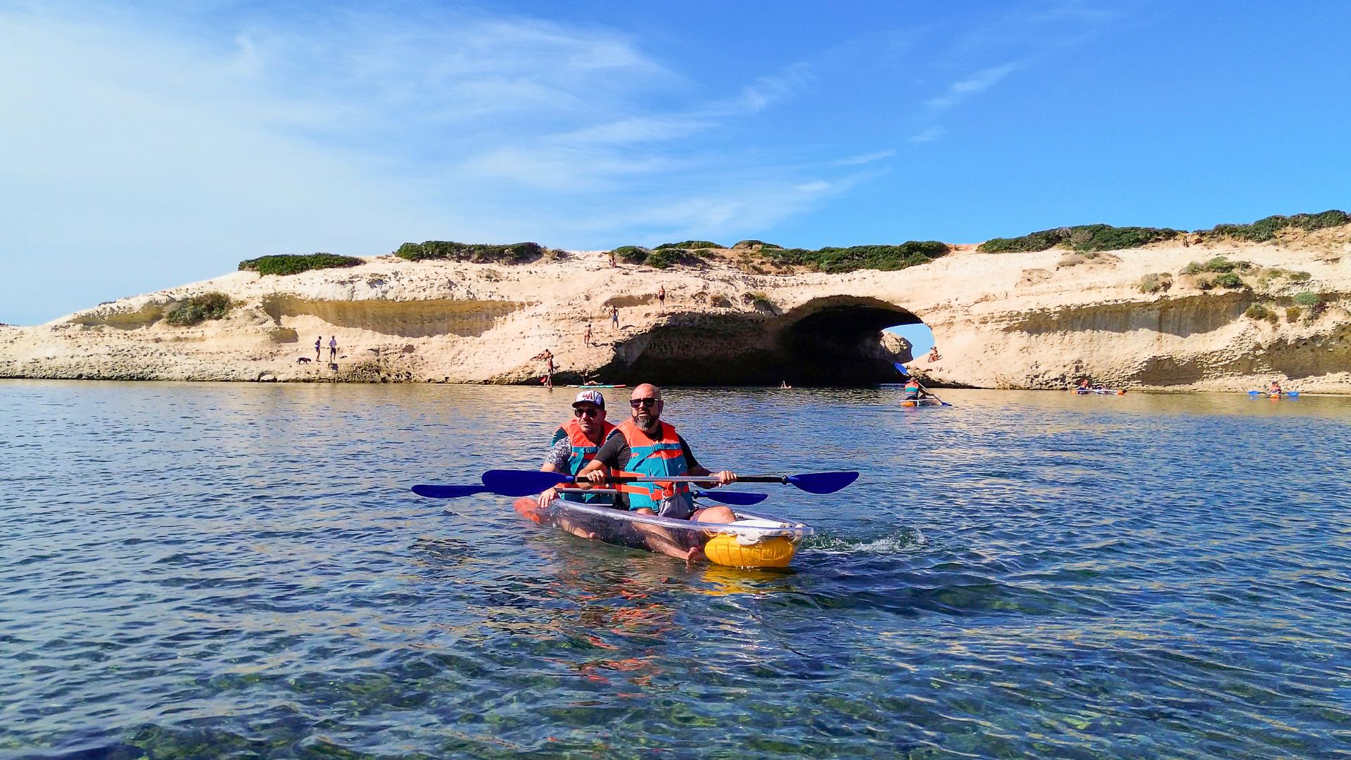 Due persone in un kayak trasparente, in acqua cristallina, vicino a una scogliera con un arco naturale