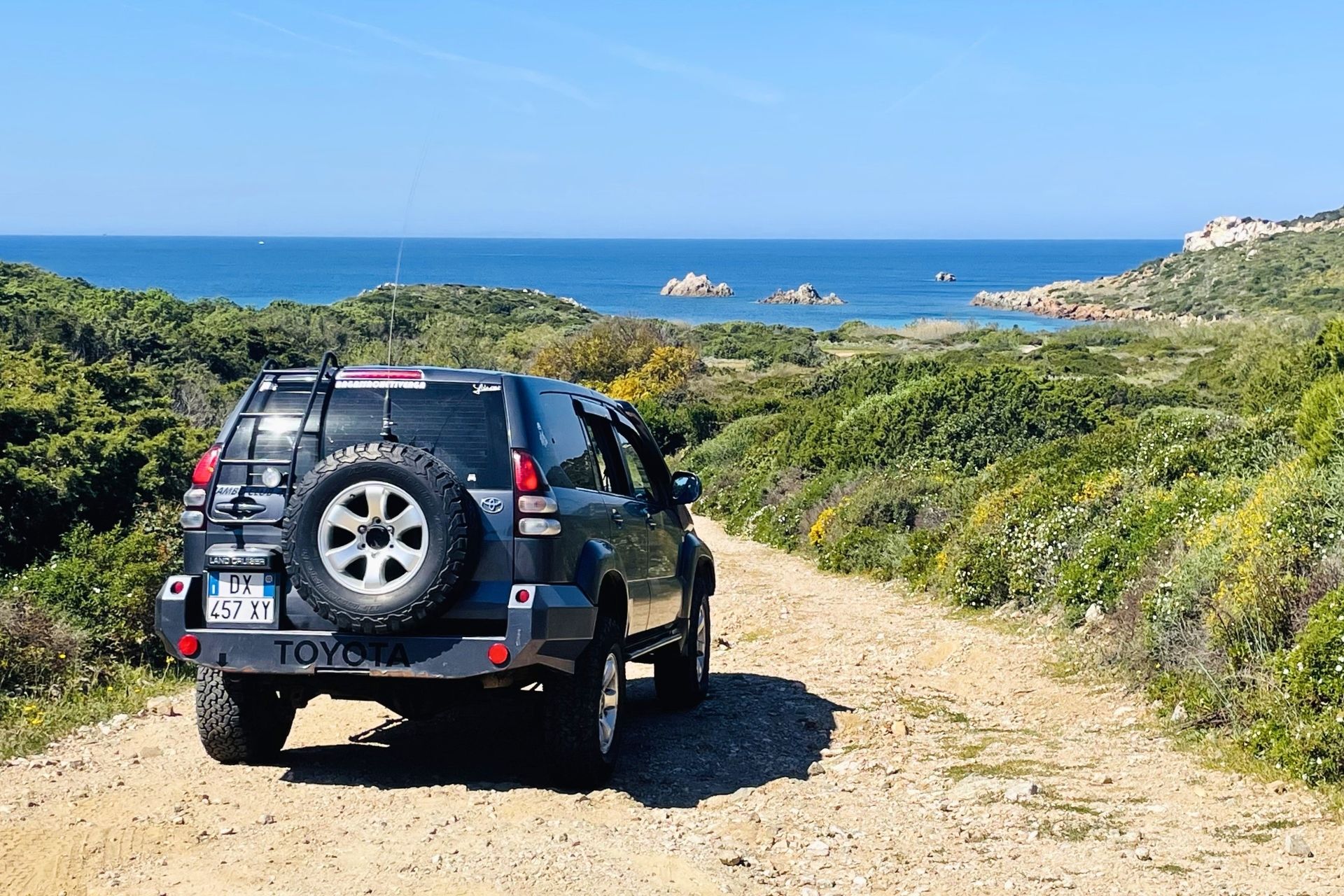 A jeep with a spare tire on the back, parked on a dirt road that gets lost in the green of a hill. In the distance, the blue sea and the rocks of the Maddalena Archipelago.