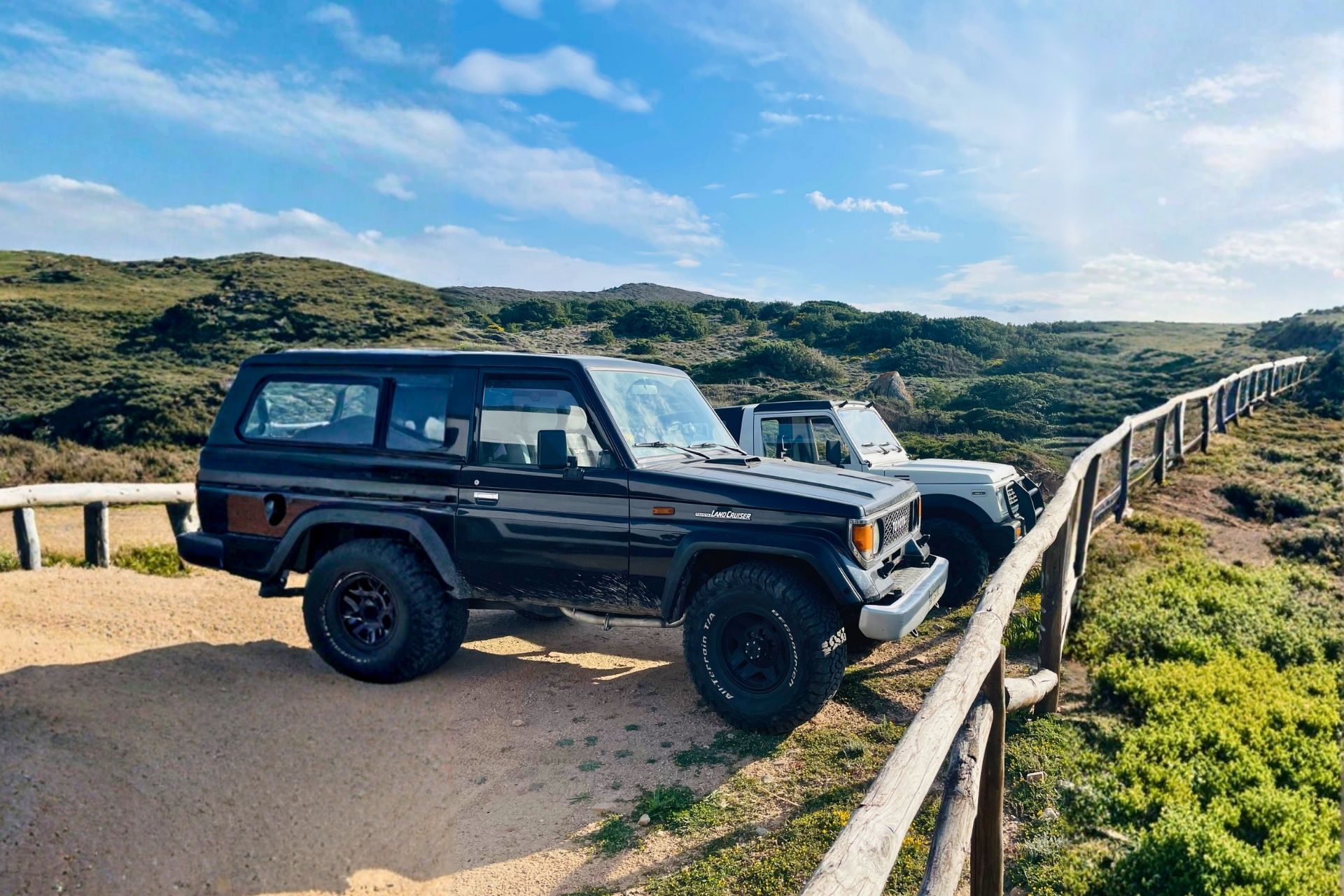 Two off-road vehicles parked on a panoramic path with hills and sea in the background