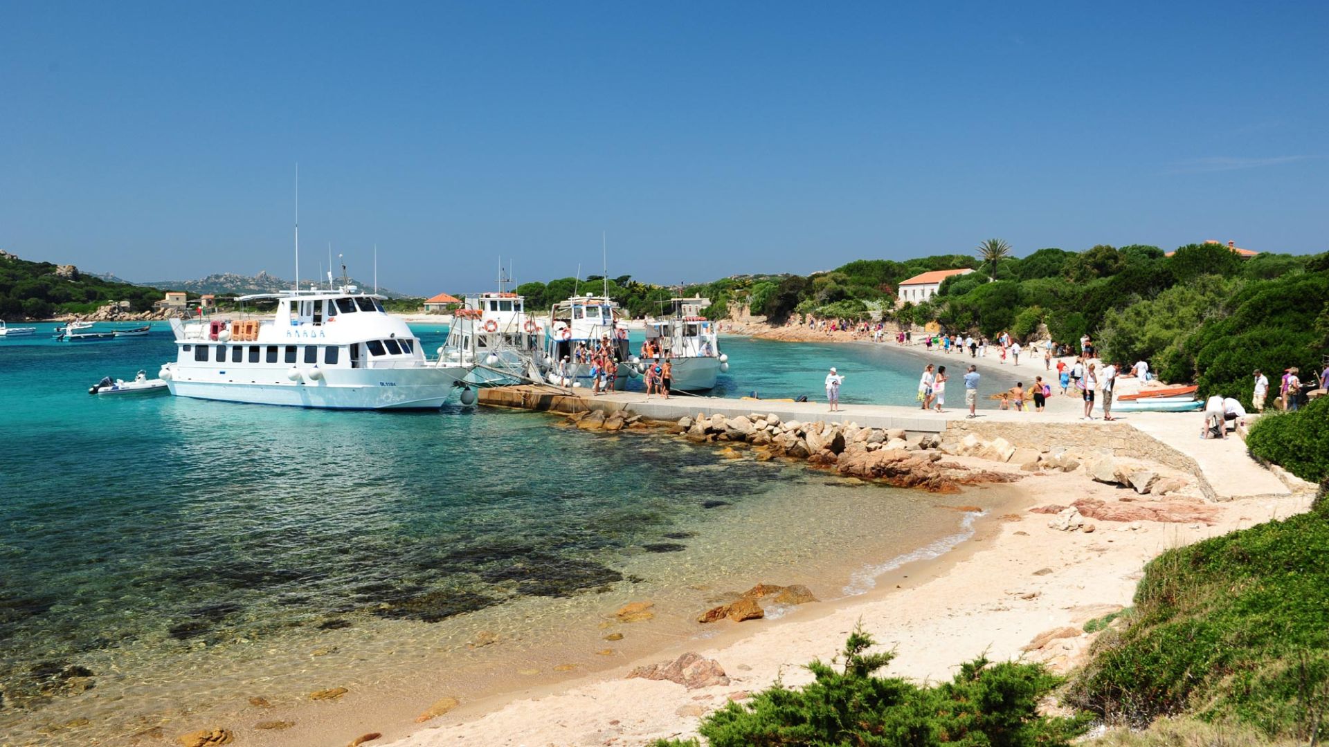 Photo d'un port avec plusieurs bateaux amarrés et des gens marchant sur la plage et sur la promenade du port