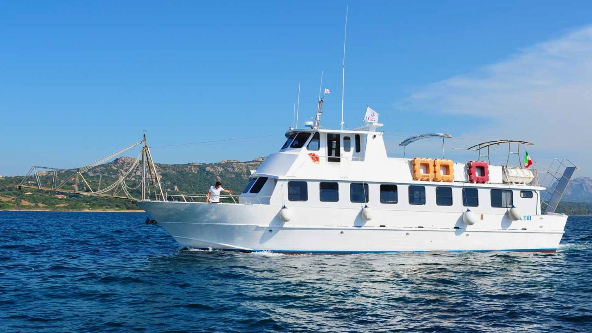 Un bateau à moteur blanc navigue sur une mer bleue avec des îles en arrière-plan. Un homme debout sur la proue tient la barre de commande. Il y a des sauveteurs, des drapeaux et une antenne à bord