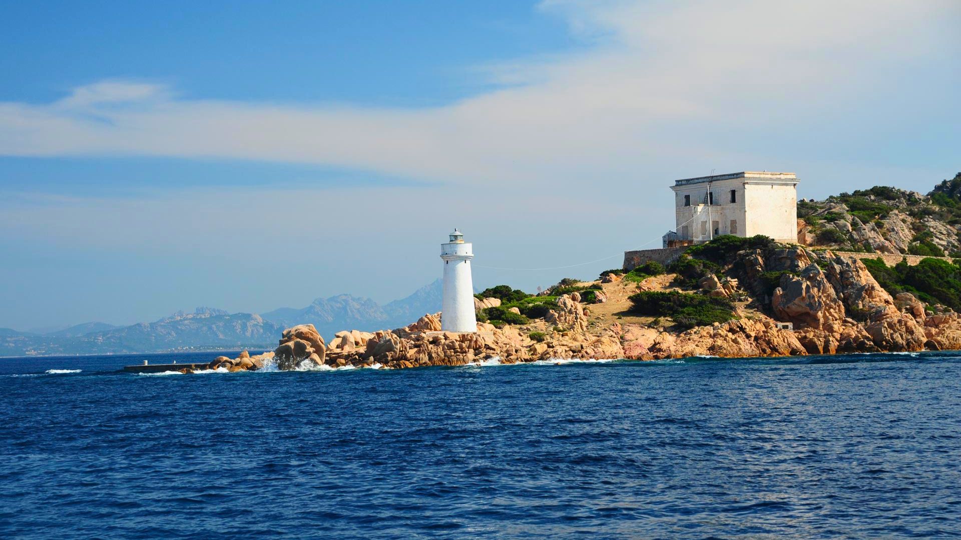 Image d'une île avec un phare et une structure blanche sur fond de mer bleue et de ciel partiellement nuageux. En arrière-plan, vous pouvez voir des montagnes et d'autres îles