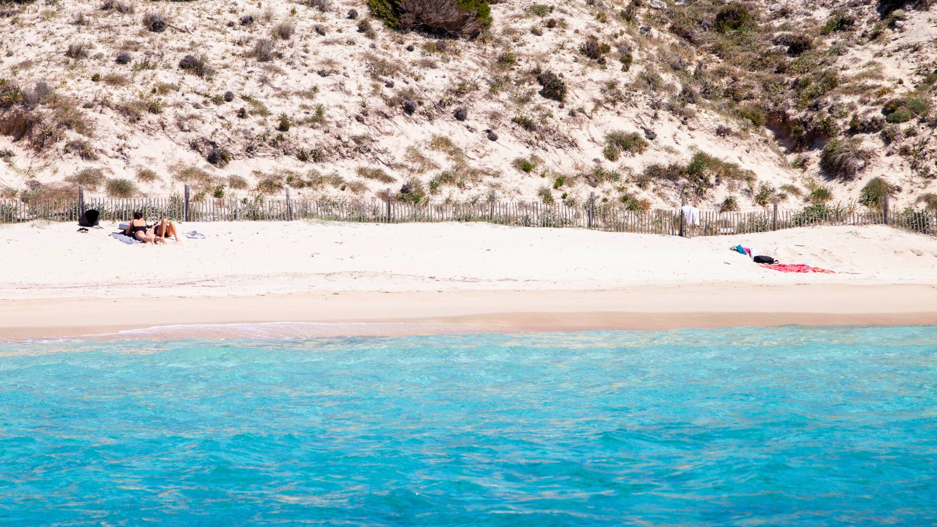 Due persone sdraiate sulla sabbia di una spiaggia con un sentiero di legno e una collina di sabbia sullo sfondo