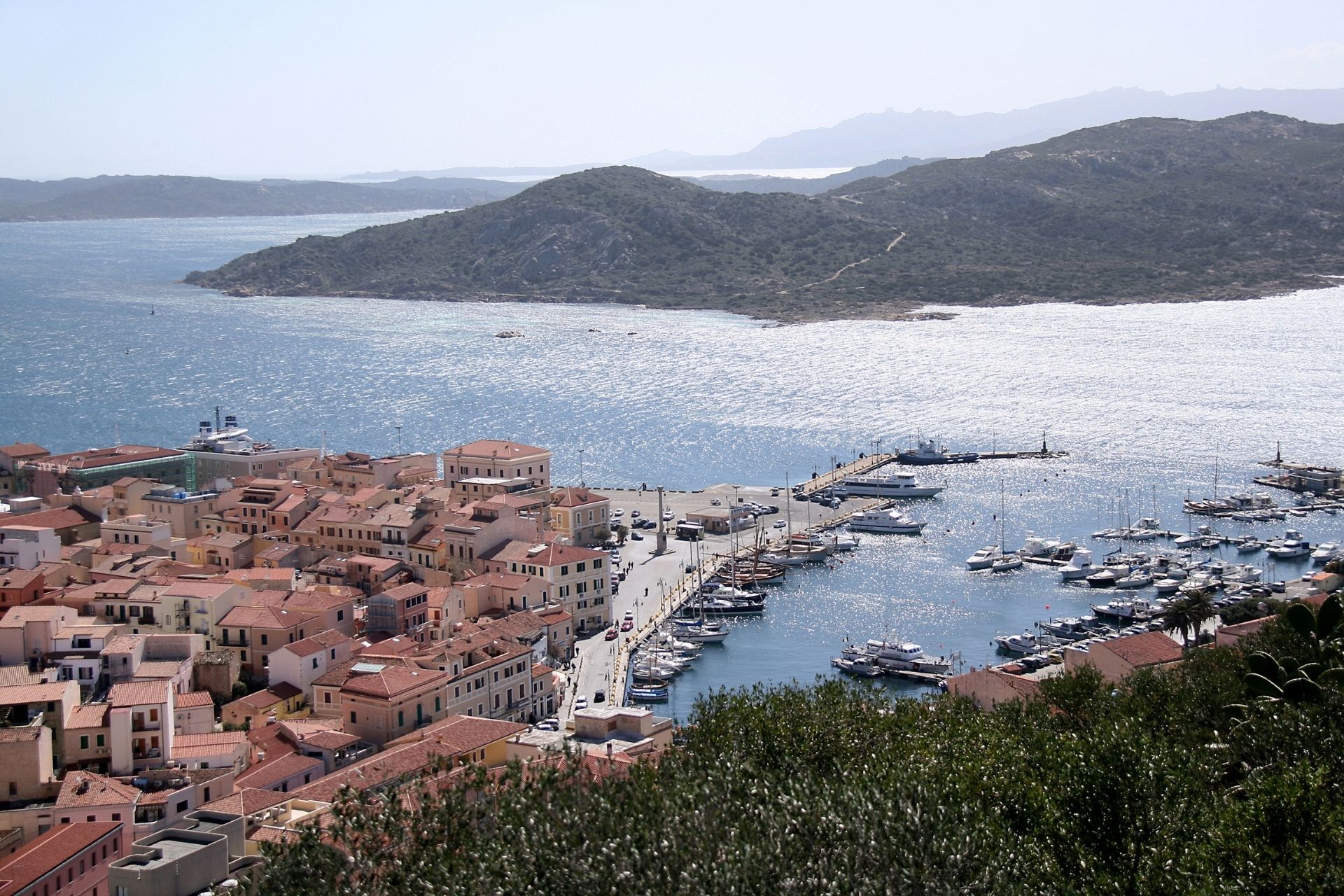 A bird's-eye view of a coastal town with a busy boat port and a street lined with red-roofed buildings.