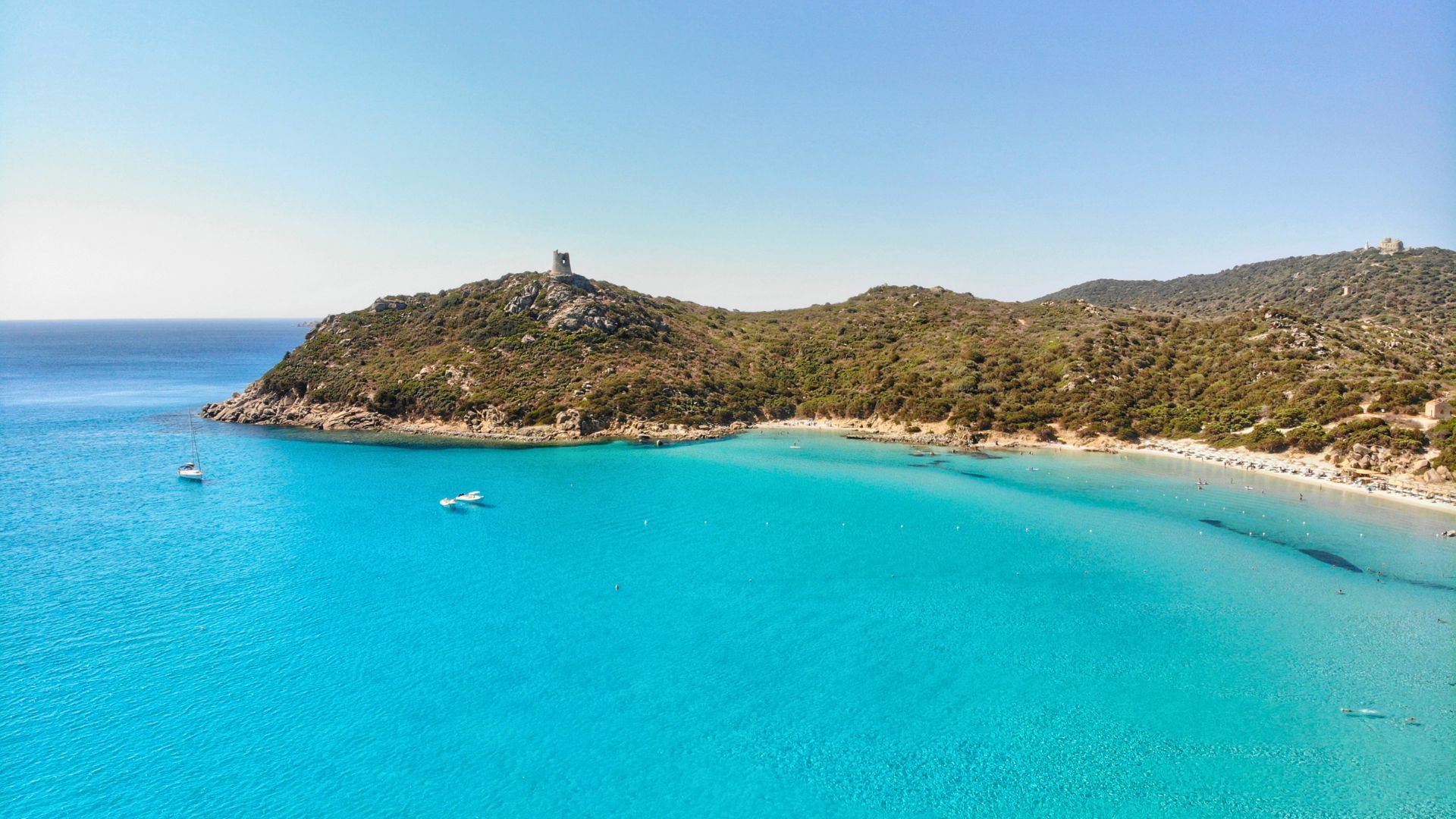 A beach with blue, verdant water, white sand and a promontory with a tower at the top