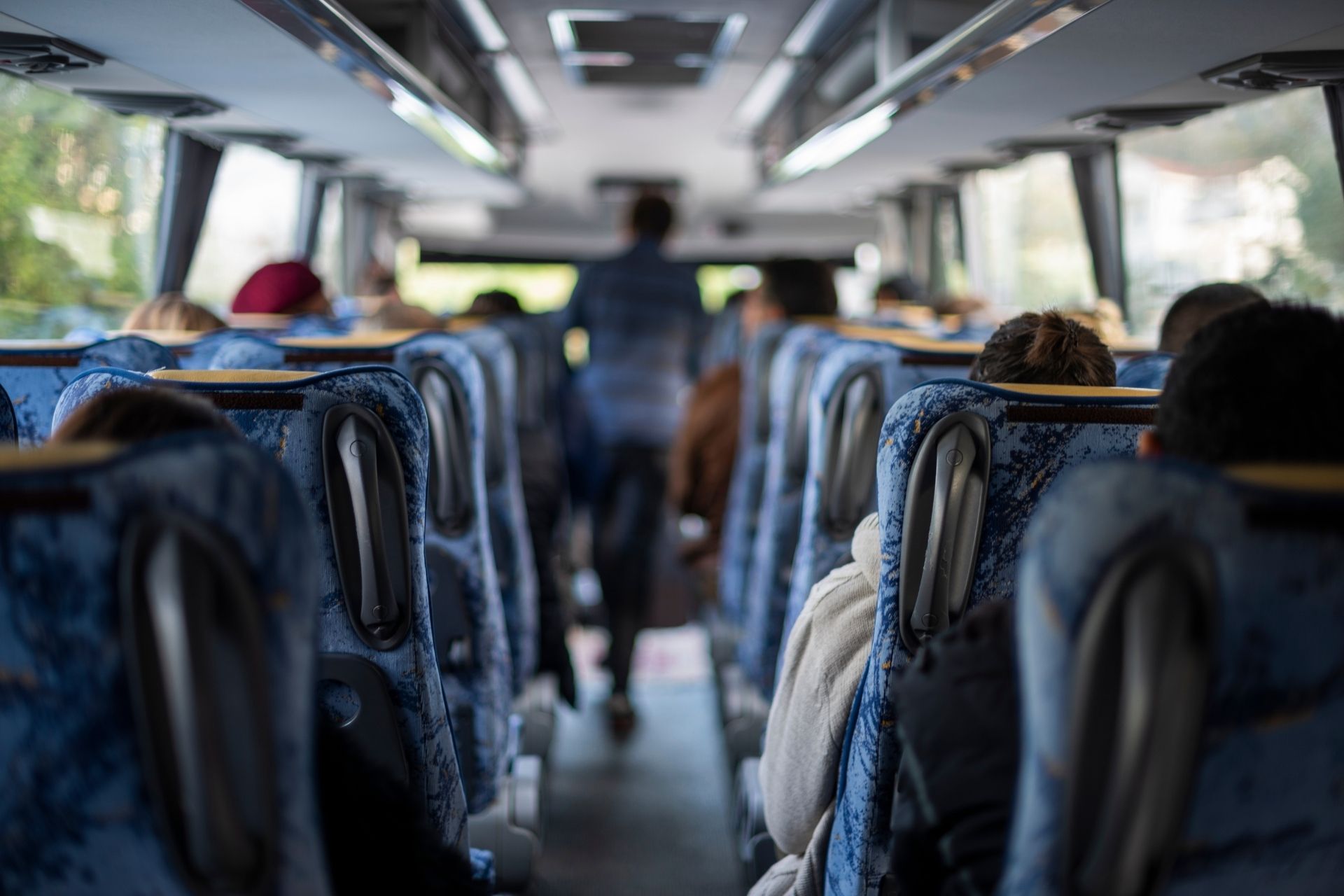 Interior of a bus with passengers seated, a man standing in front of them, side windows and roof with lights.