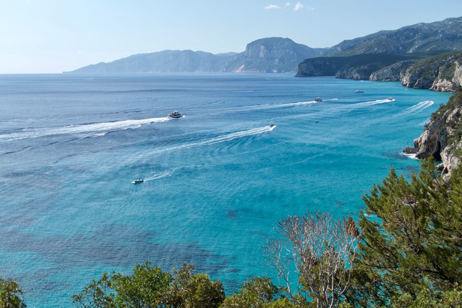 A group of boats sail in an intense blue sea with mountains in the background and a rocky coastline.