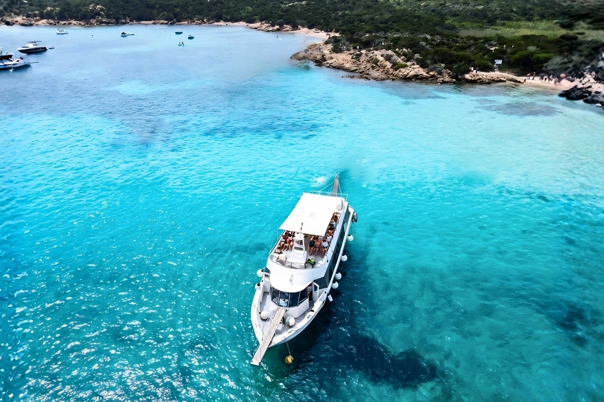 Una motonave naviga in acque turchesi, con alcune persone a bordo, vicino a una spiaggia con rocce e vegetazione verde. Sullo sfondo, altre imbarcazioni e un paesaggio naturale con vegetazione e rocce.