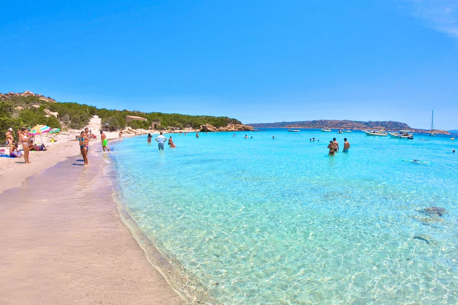 Persone che nuotano e prendono il sole su una spiaggia con acqua cristallina e sabbia bianca, circondata da isole e vegetazione. Un'esperienza di relax e natura nel nord della Sardegna.
