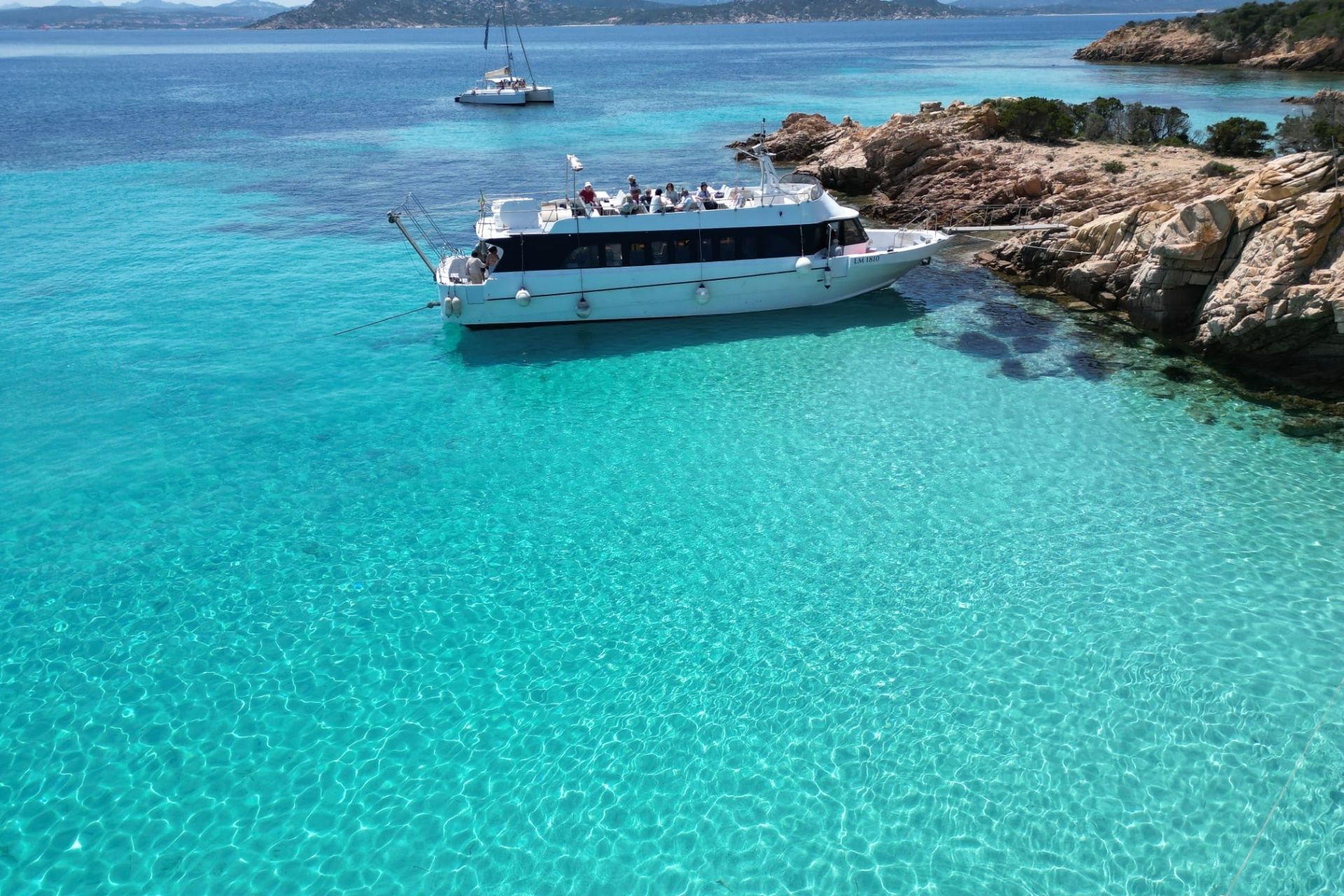 Immagine di una motonave ormeggiata in acque turchesi, con un'isola rocciosa sullo sfondo e un'altra barca in lontananza.