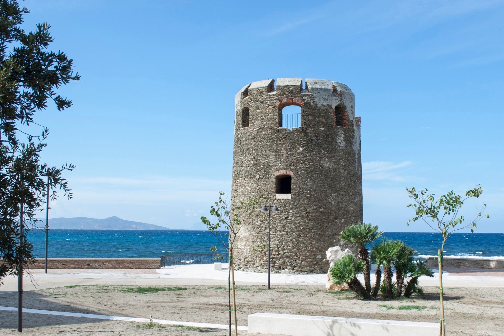 Coastal tower with windows and battlements, surrounded by trees and plants, with a view of the sea and mountains in the background.