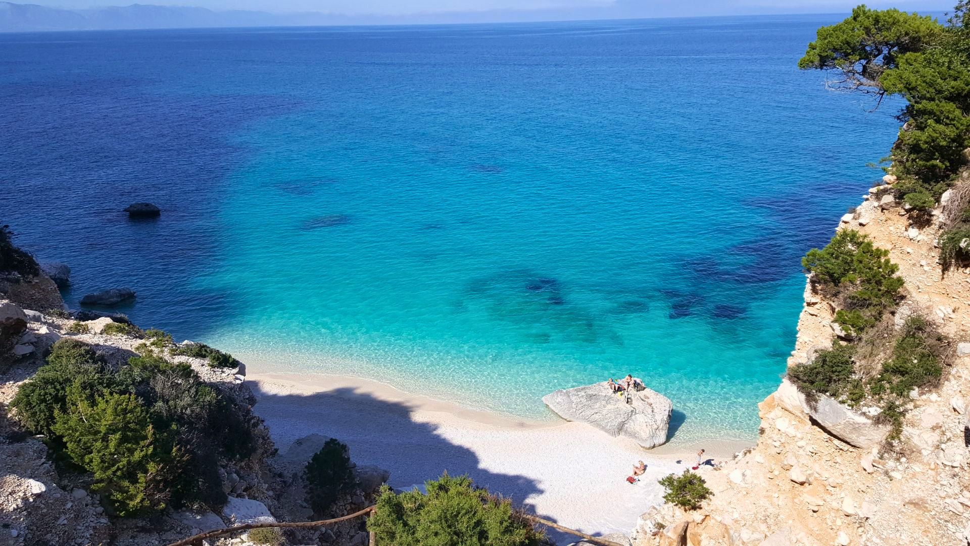Ein Motorschiff segelt in einem Golf mit türkisfarbenem Wasser und einem weißen Strand, mit Menschen, die auf einem Felsen sitzen und Bäume und Felsen an der Seite einer Klippe