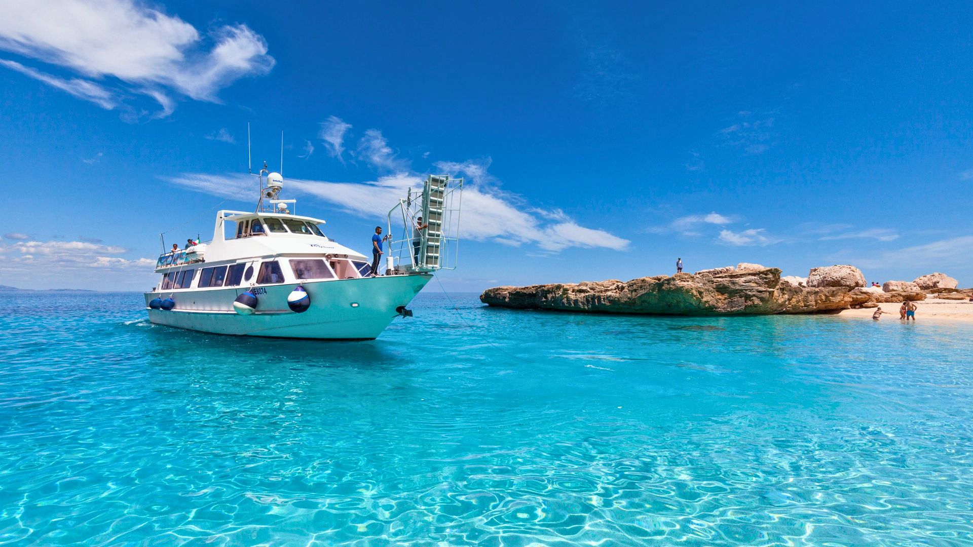 Ein Motorschiff segelt in kristallklarem Wasser mit einer Küste mit Klippen und einer kleinen Insel im Hintergrund. Es gibt Leute auf dem Boot und andere an der Küste