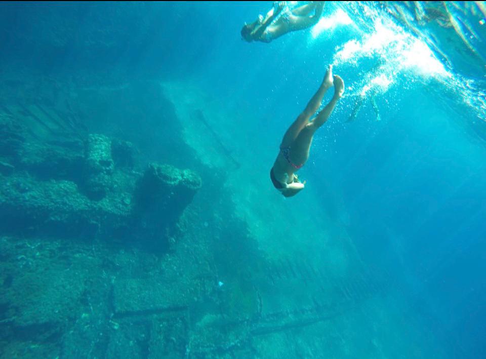 Dos personas nadando en el mar, cerca de un acantilado y un naufragio, con aguas cristalinas y azules.
