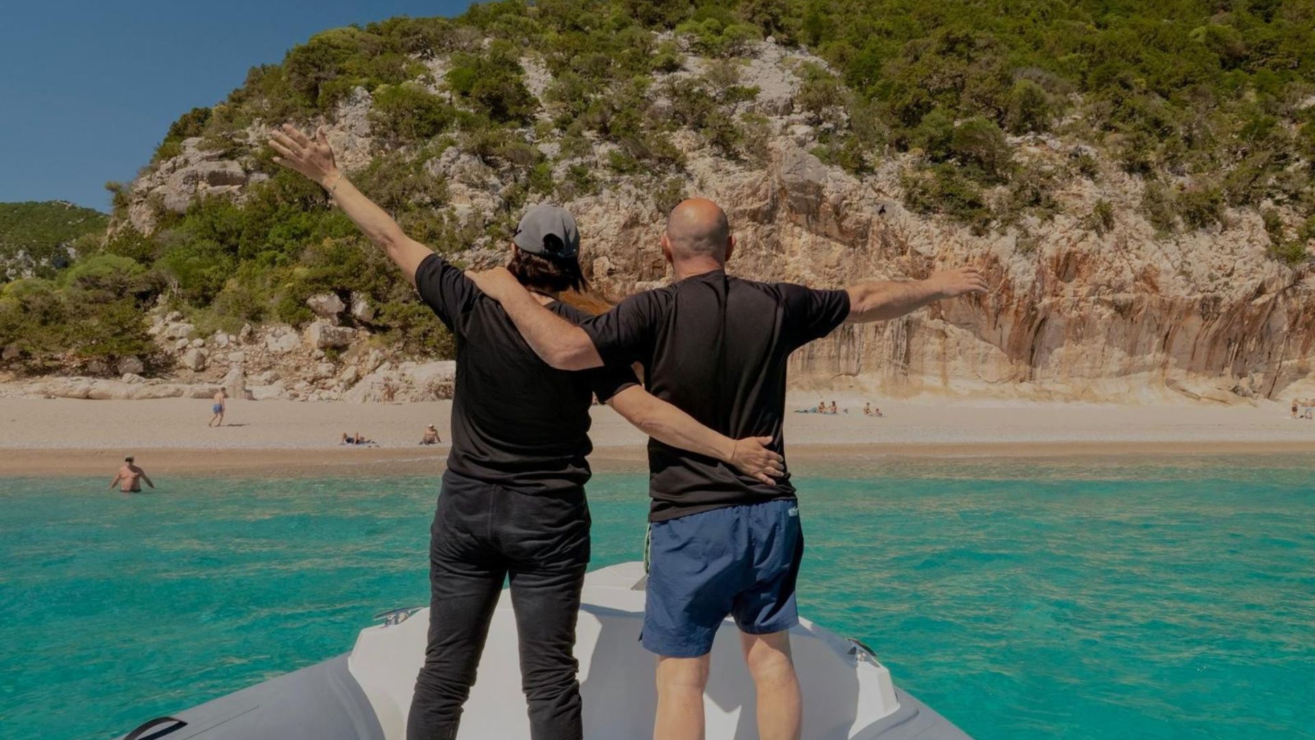Two men standing on a dinghy with their arms raised in front of a beach and a crystal clear sea. The text talks about a dinghy tour in the Gulf of Orosei with an aperitif
