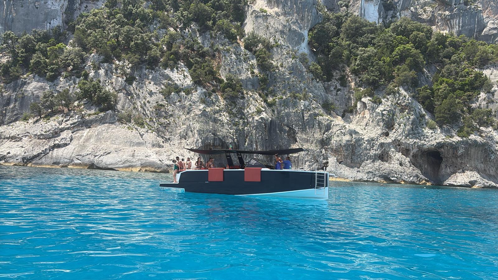 A group of people on a mini yacht sail in crystal clear waters near a rocky cliff with vegetation.