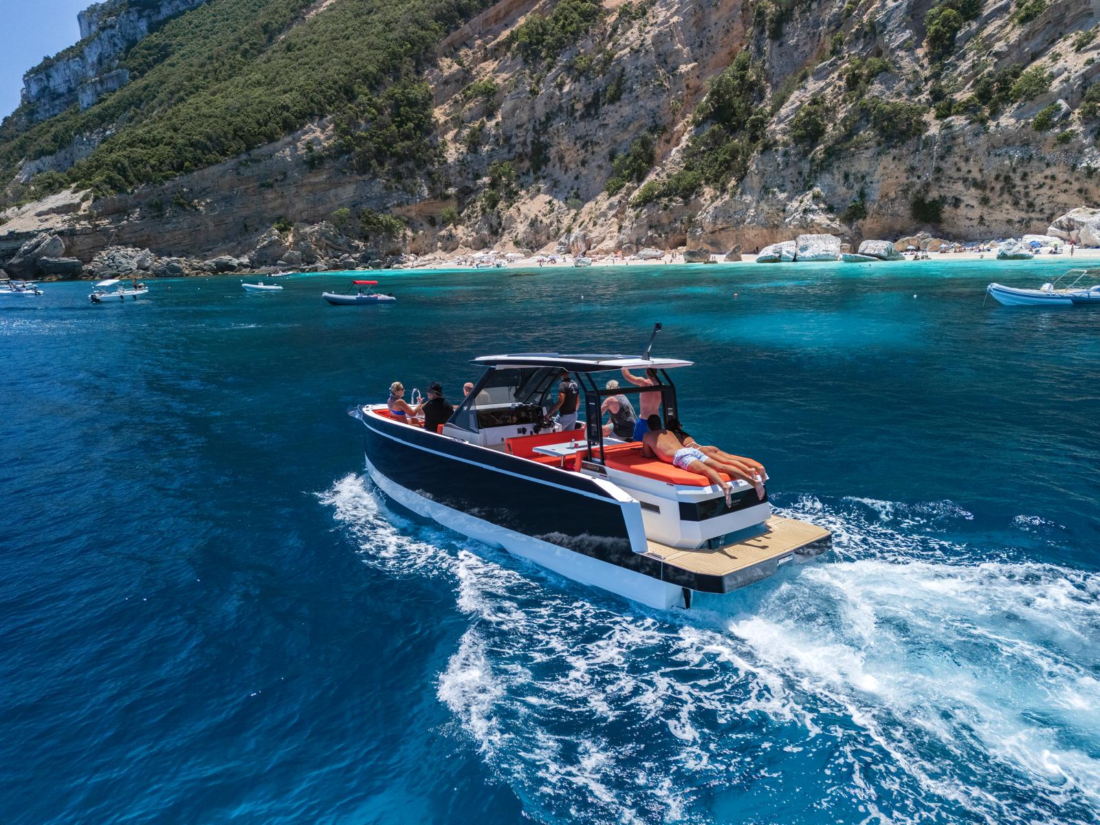 A group of people on a mini yacht sail in crystal clear waters with turquoise shades, near cliffs and coves accessible only by sea.