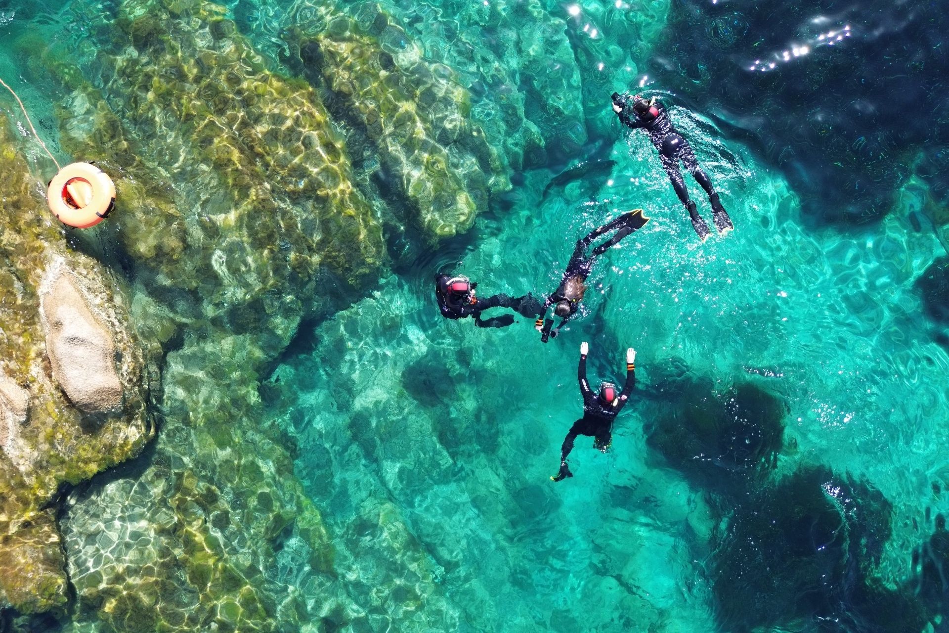 Imagen de tres personas sumergidas en las aguas cristalinas de Tavolara y Molara, con rocas y un salvavidas naranja a la vista.