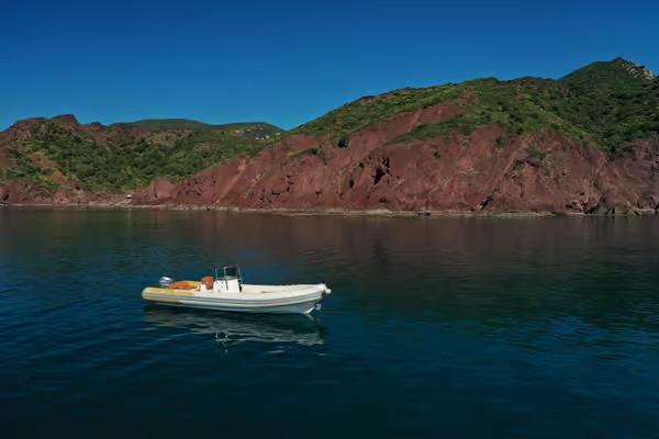 Immagine di un gommone in acqua con uno sfondo di scogliere e montagne colorate. L'acqua e' limpida e trasparente.