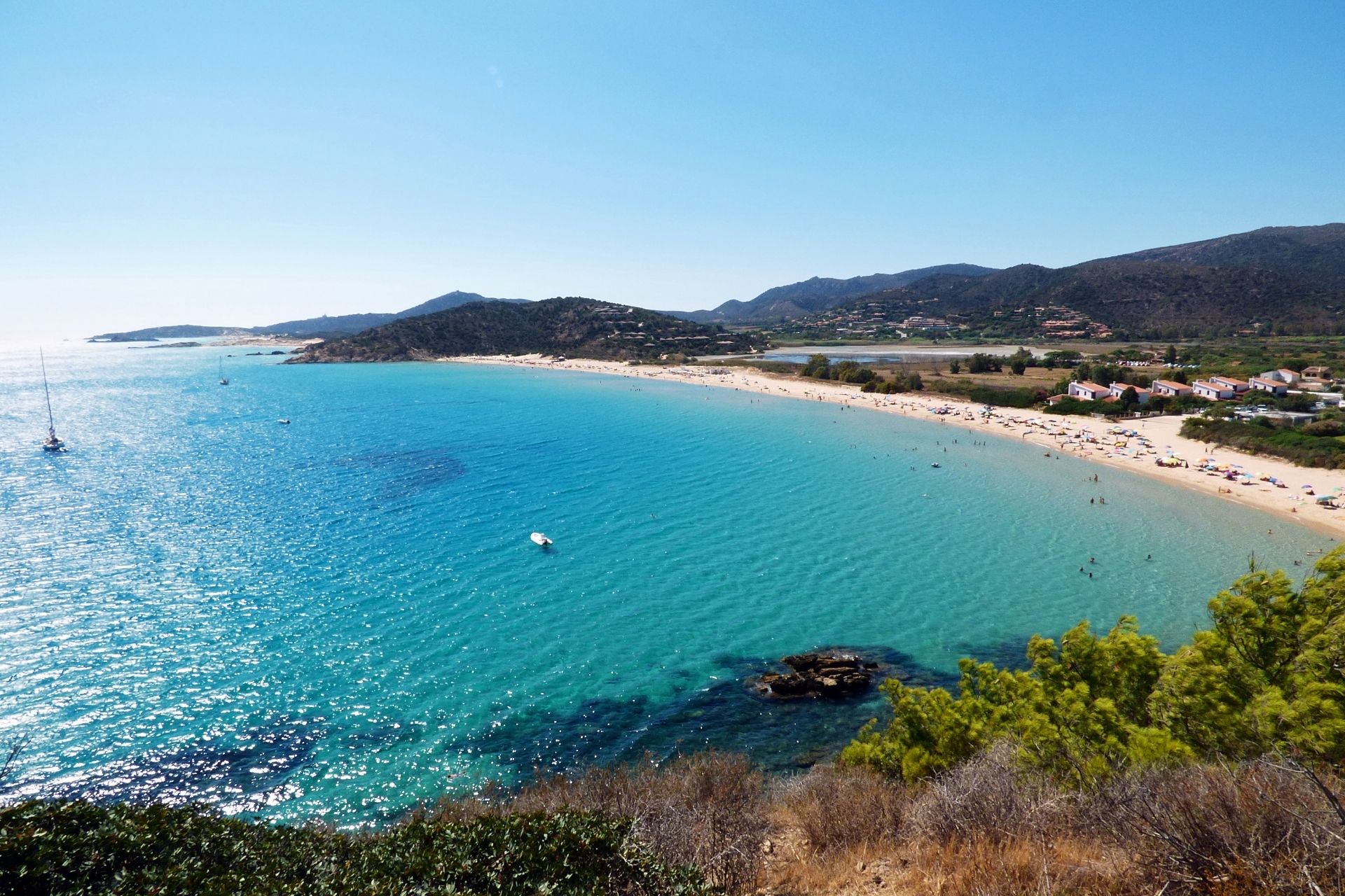 Ein Strand mit blauem Wasser und weißem Sand, umgeben von Hügeln und grüner Vegetation, mit einigen Segelbooten in der Ferne.