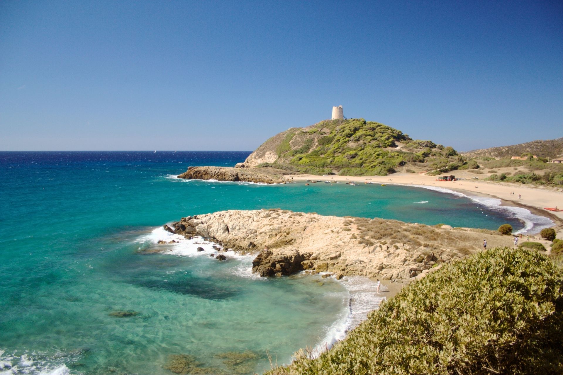 Ein Bild von einem Strand mit einem Leuchtturm im Hintergrund, einem blauen Meer und einer felsigen Küste mit grüner Vegetation. Eine kleine Gruppe von Menschen geht am Strand spazieren. Im Hintergrund eine Insel mit einem Leuchtturm. Im Hintergrund ein ruhiges, blaues Meer. Der Himmel ist klar und blau.