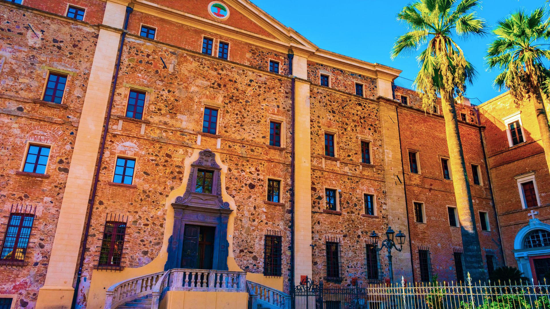 Edificio storico con facciata in pietra e finestre ad arco, con palme davanti e una scala esterna