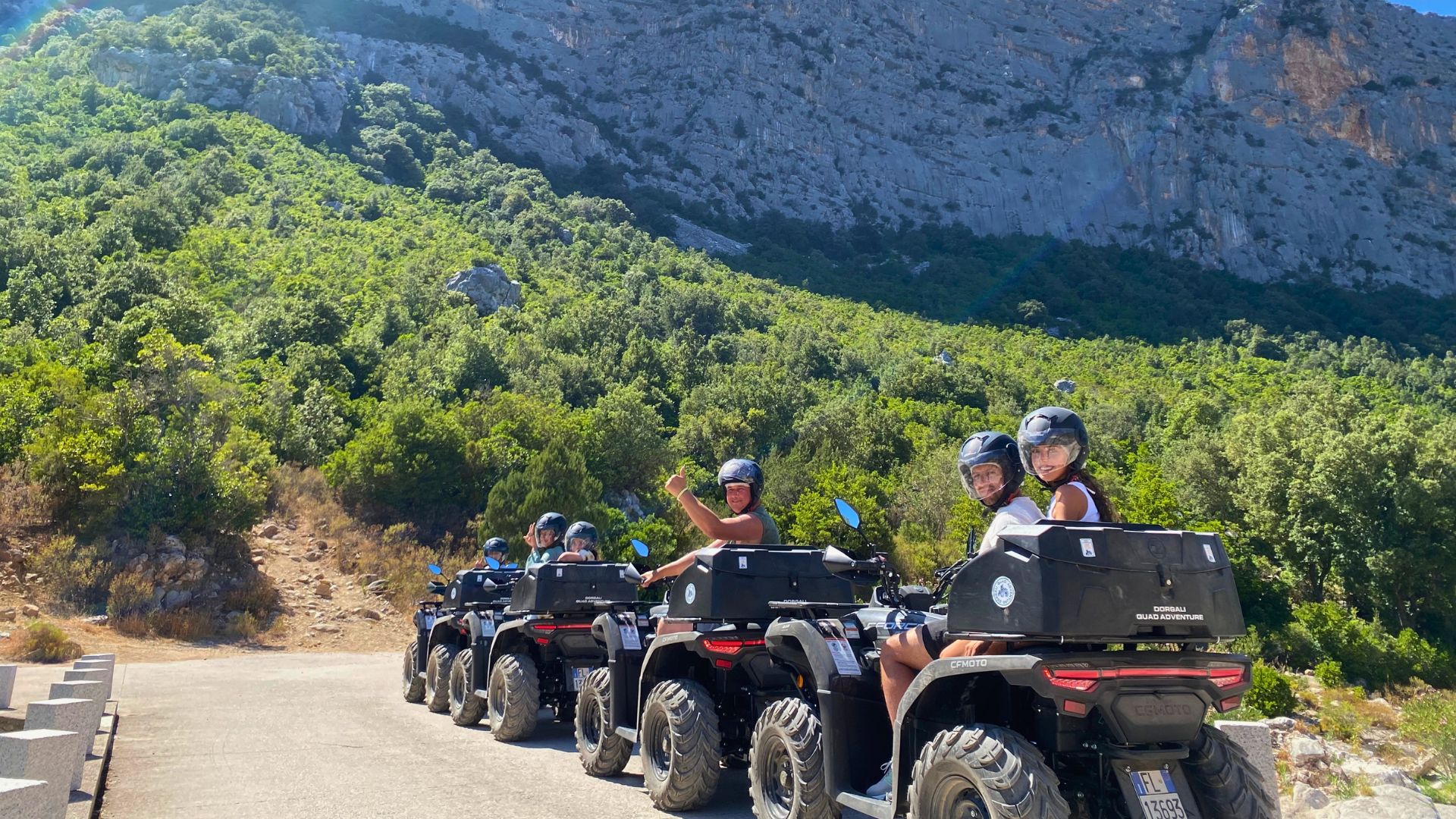 Gruppo di persone su quad in una strada sterrata con paesaggio di montagna e mare sullo sfondo
