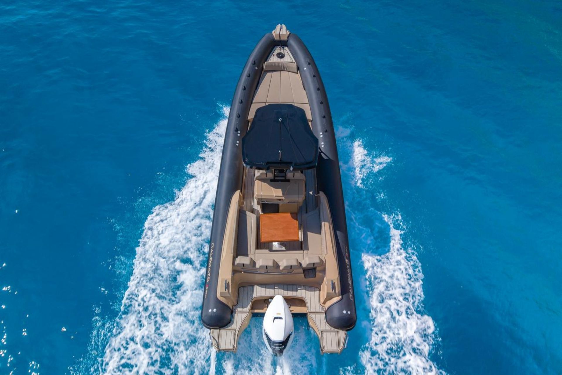 Image d'un dériveur au toit noir naviguant en pleine mer avec le golfe d'Orosei en arrière-plan en Sardaigne. Le canot dispose d'un coin repas couvert avec table et chaises et d'un espace détente avec canapés et coussins. La mer est calme et le ciel est clair.