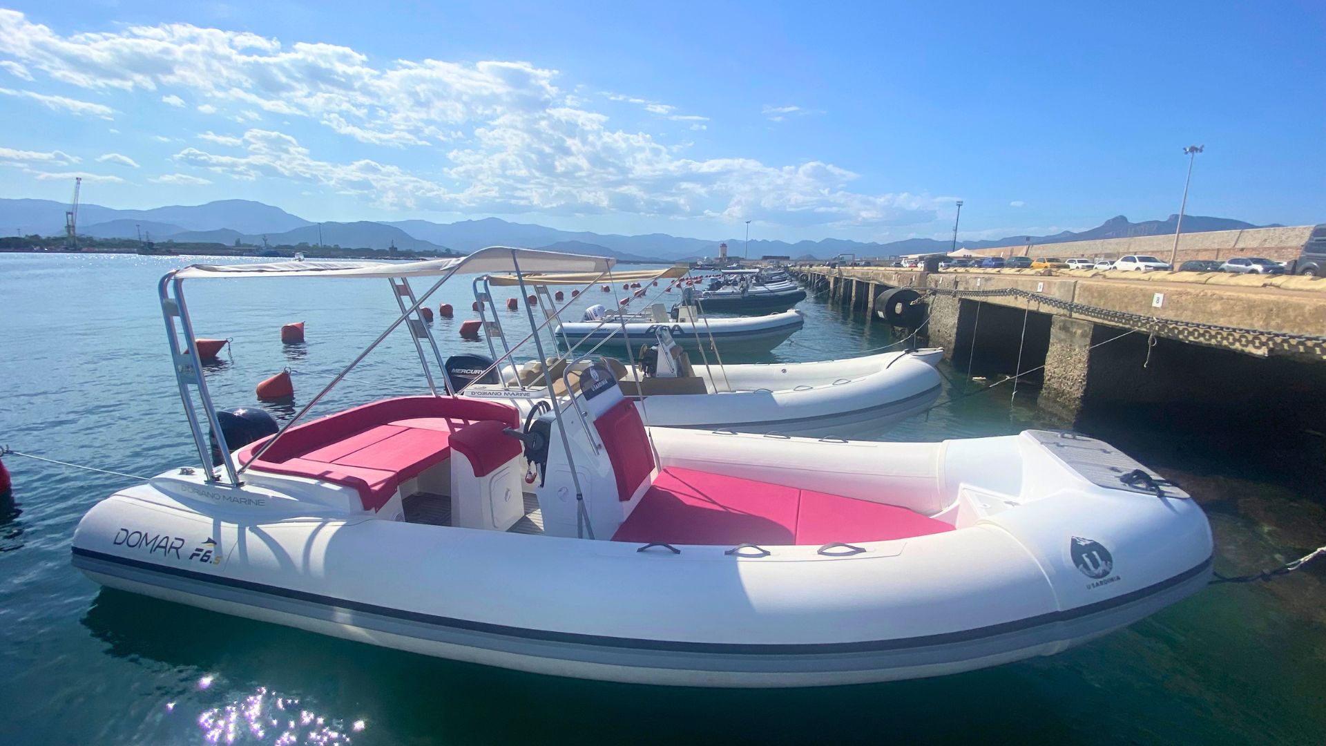 Image of a white and red dinghy moored at the port with beaches and mountains in the background.