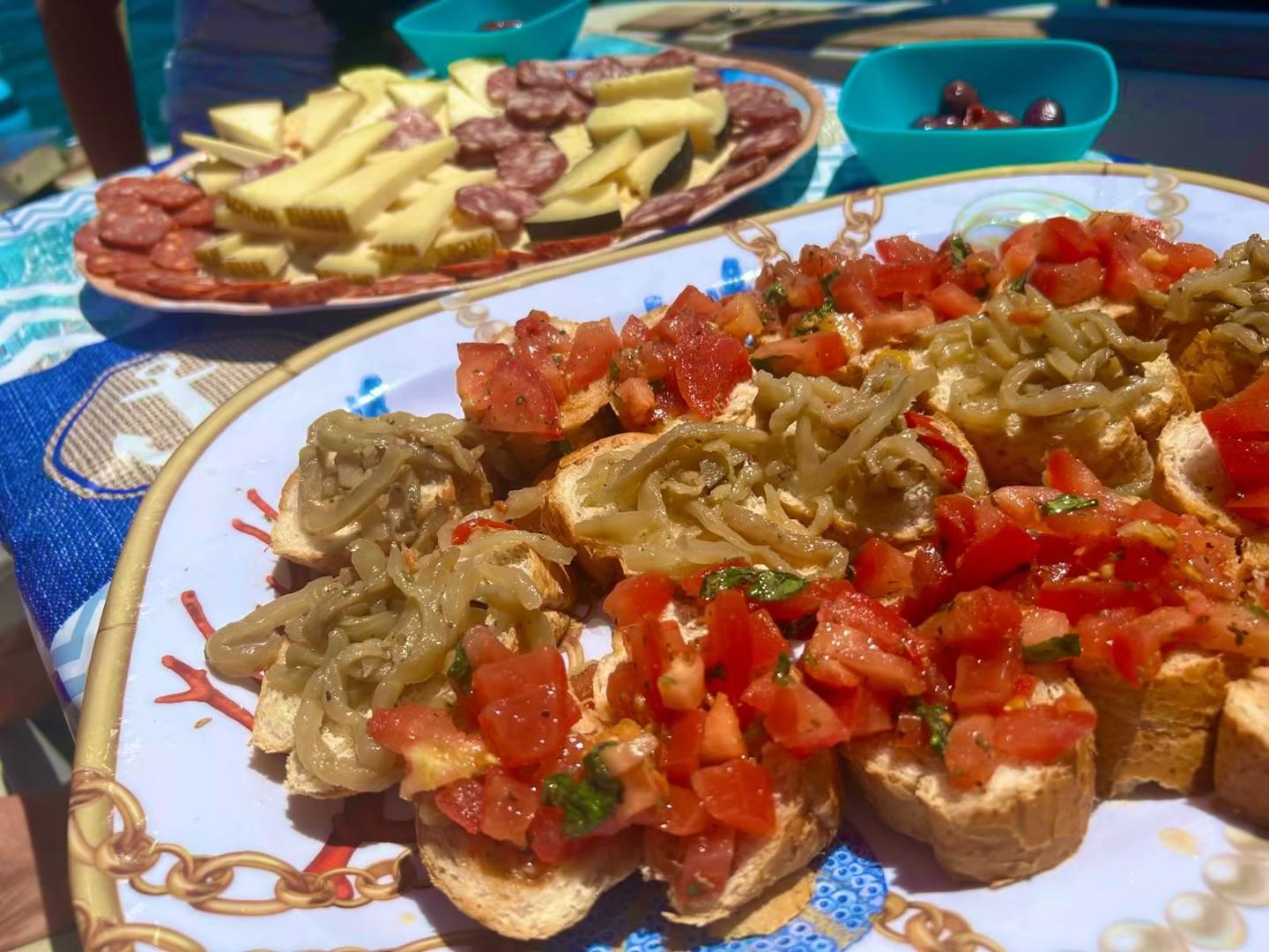 Two plates of food on a table overlooking the sea, one with bread and vegetables, the other with cold cuts and cheeses.