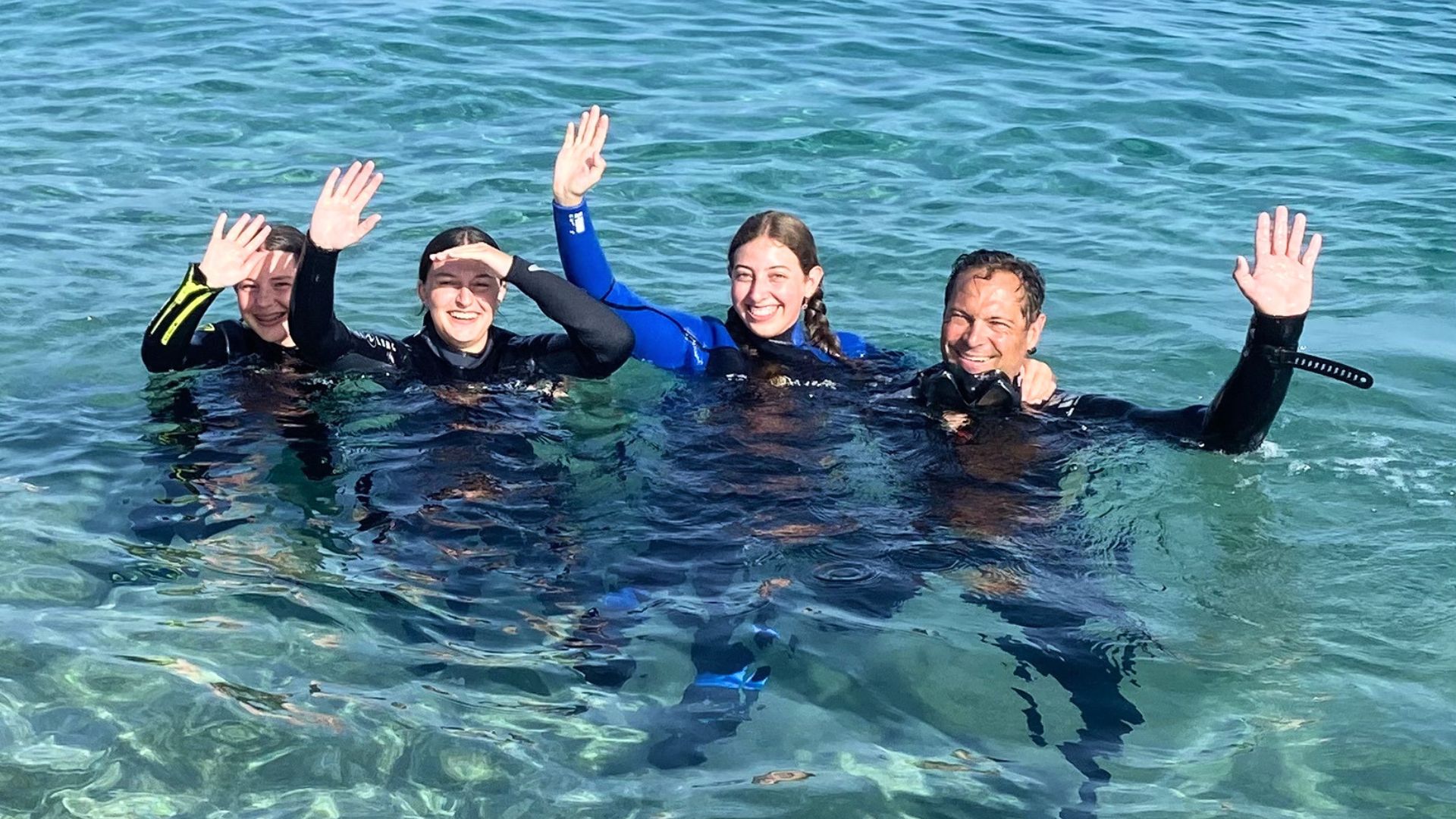 Grupo de personas en trajes de baño y trajes de buceo, sonriendo y en el agua, con las manos levantadas