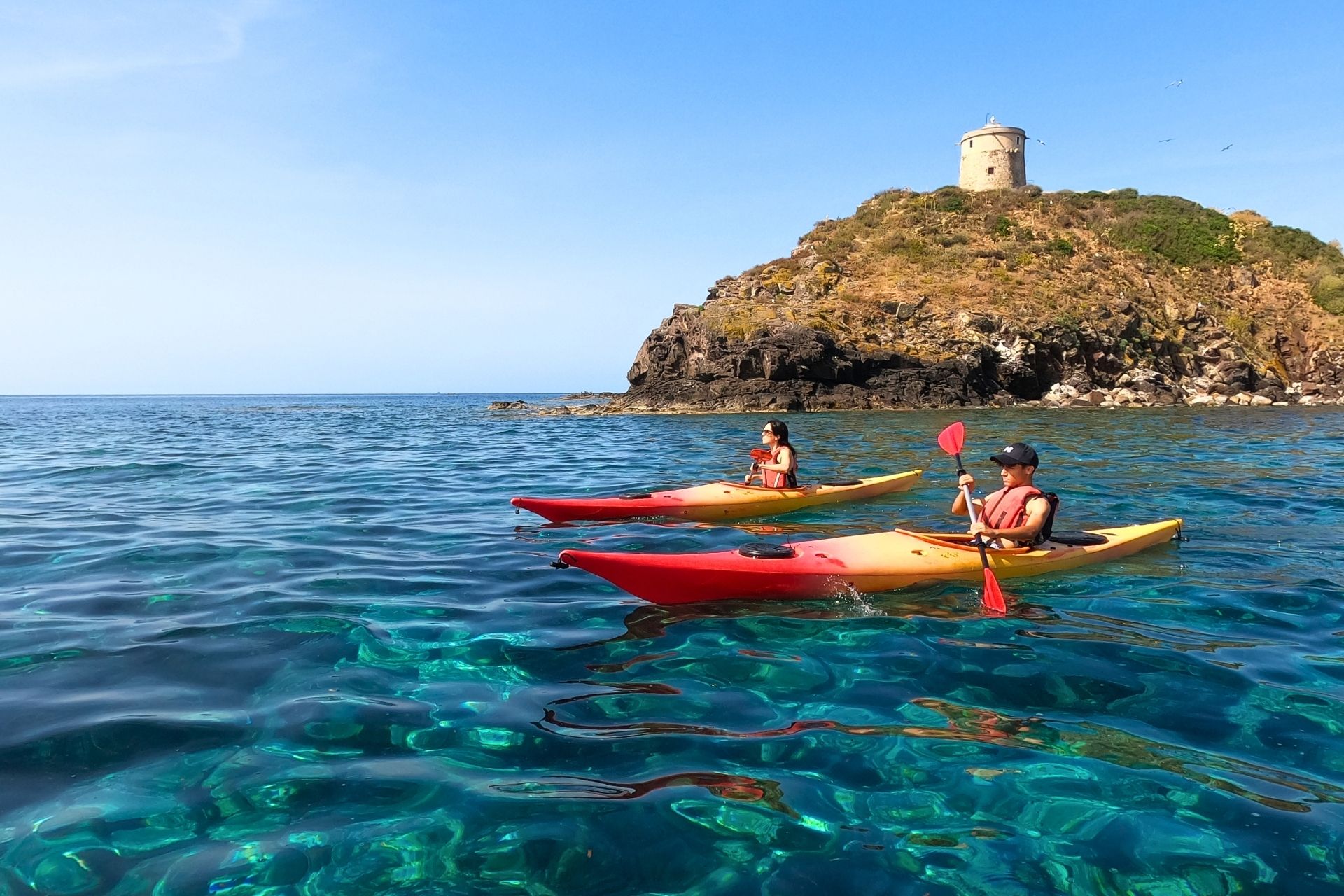 Two people in red and yellow kayaks near an island with a tower on the top, a clear blue sea in the background.