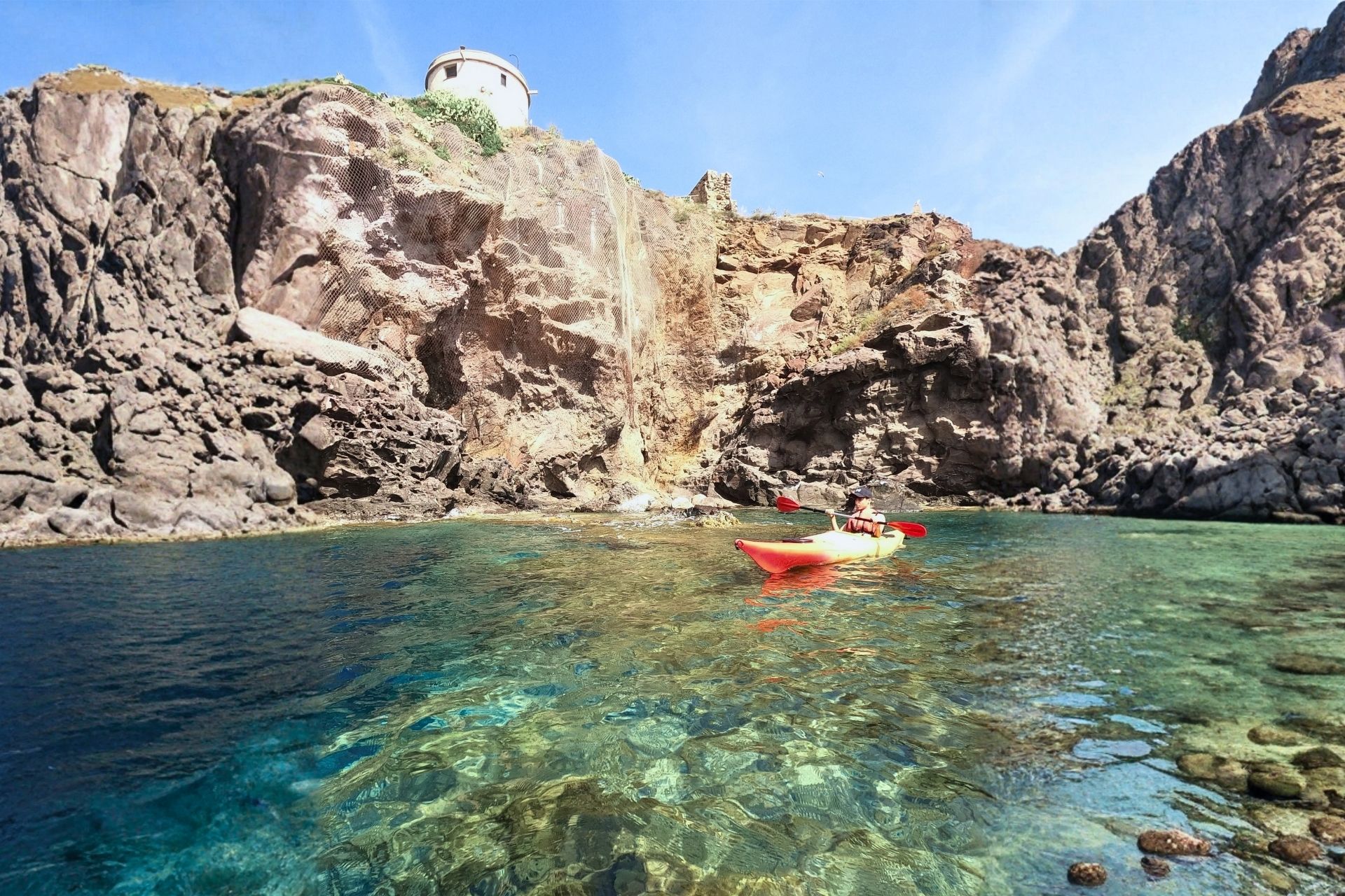 A kayaker in technical clothing sails in crystal clear waters in front of rocky cliffs. The kayaker wears a hat and a diving suit. In the background a small white building.