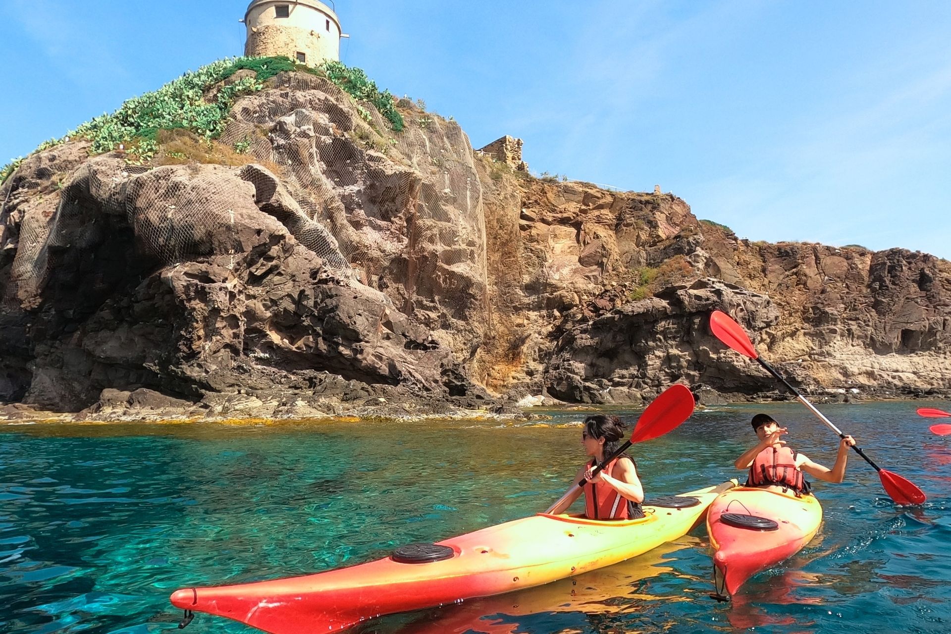Two people in red kayaks near a rocky cliff, with a lighthouse in the background and crystal clear waters.