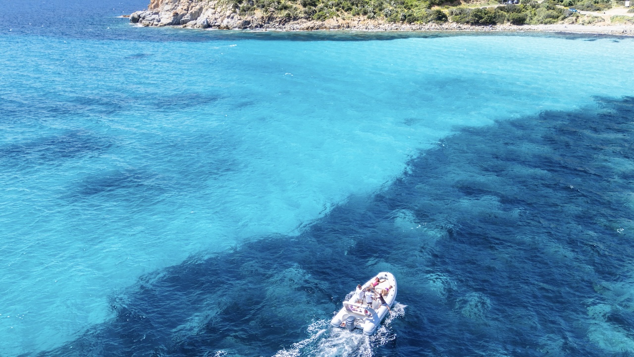 Una barca a vela con persone a bordo naviga in mare aperto con acque cristalline e una costa rocciosa sullo sfondo