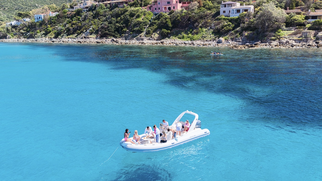 Un gruppo di persone su una barca a vela con alle spalle una costa rocciosa e case in lontananza