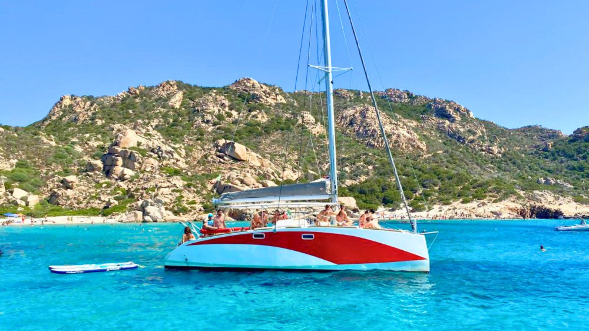Catamaran à voile naviguant dans une mer cristalline avec des passagers à bord et des montagnes en arrière-plan