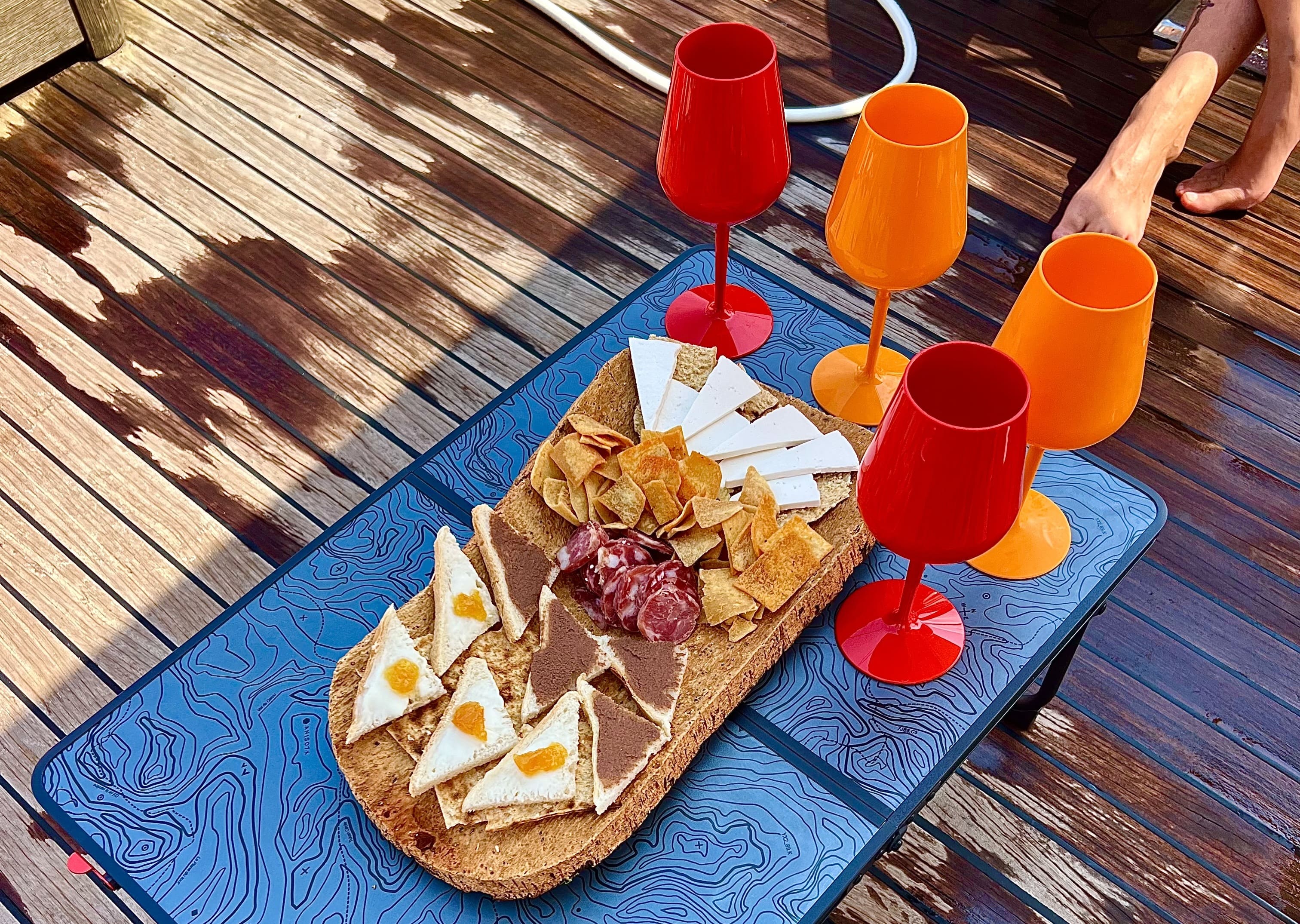 Mesa con aperitivos, embutidos y quesos en un barco con vasos de colores. Al fondo, una persona tumbada en una terraza.