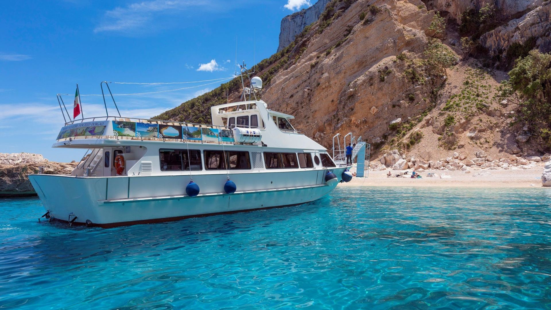 Bild eines weißen Motorschiffs, das in kristallklarem Wasser in der Nähe einer felsigen Küste mit mediterraner Vegetation vor Anker liegt