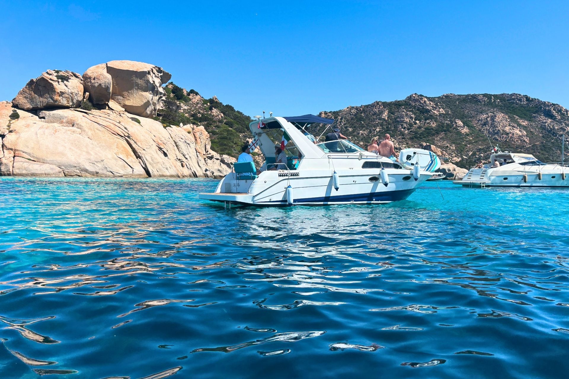 A sailboat with some passengers sails through crystal clear waters and granite cliffs in the archipelago of La Maddalena.