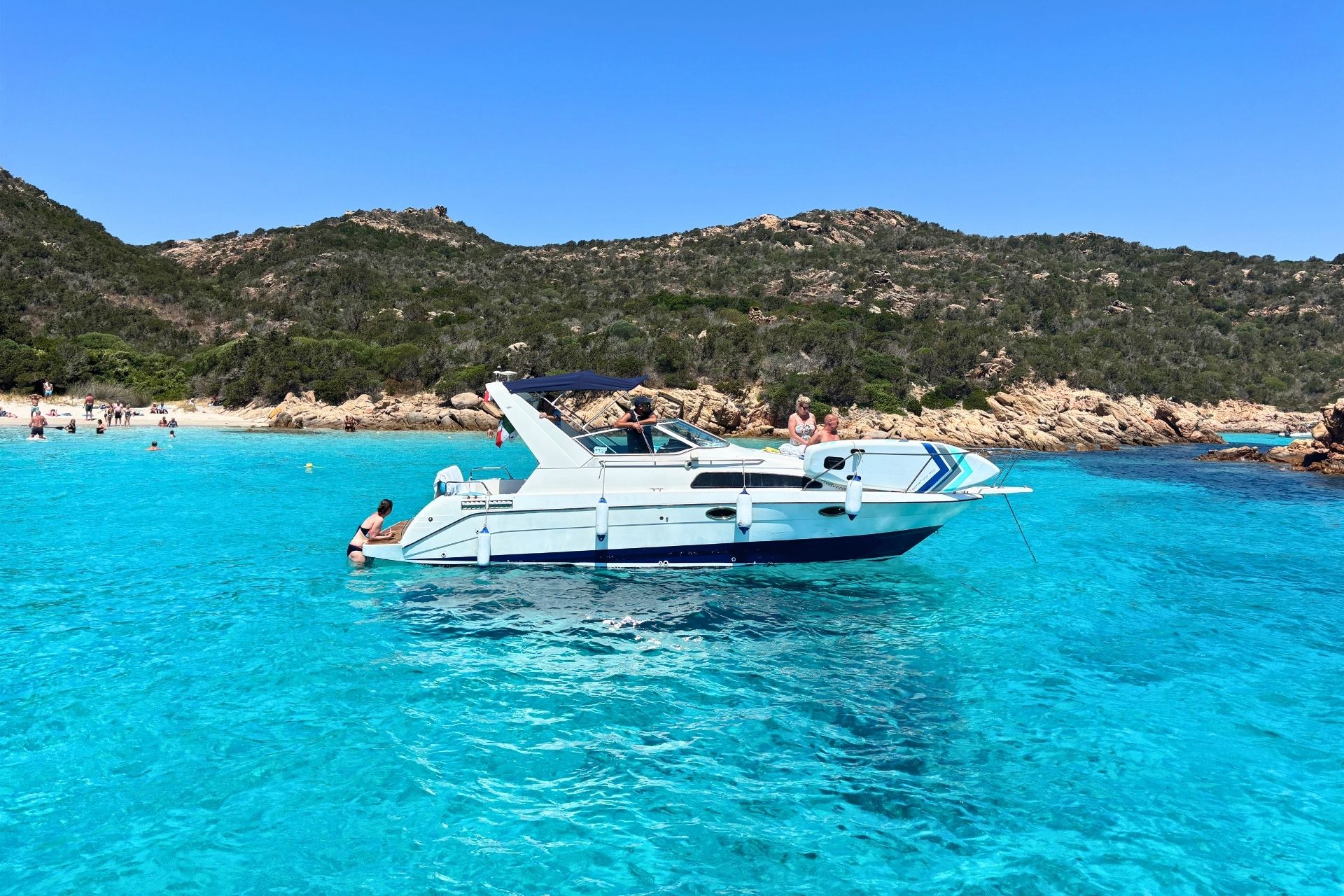 Image of a motorboat in the water with people swimming and lying on the beach, mountains and rocks in the background.