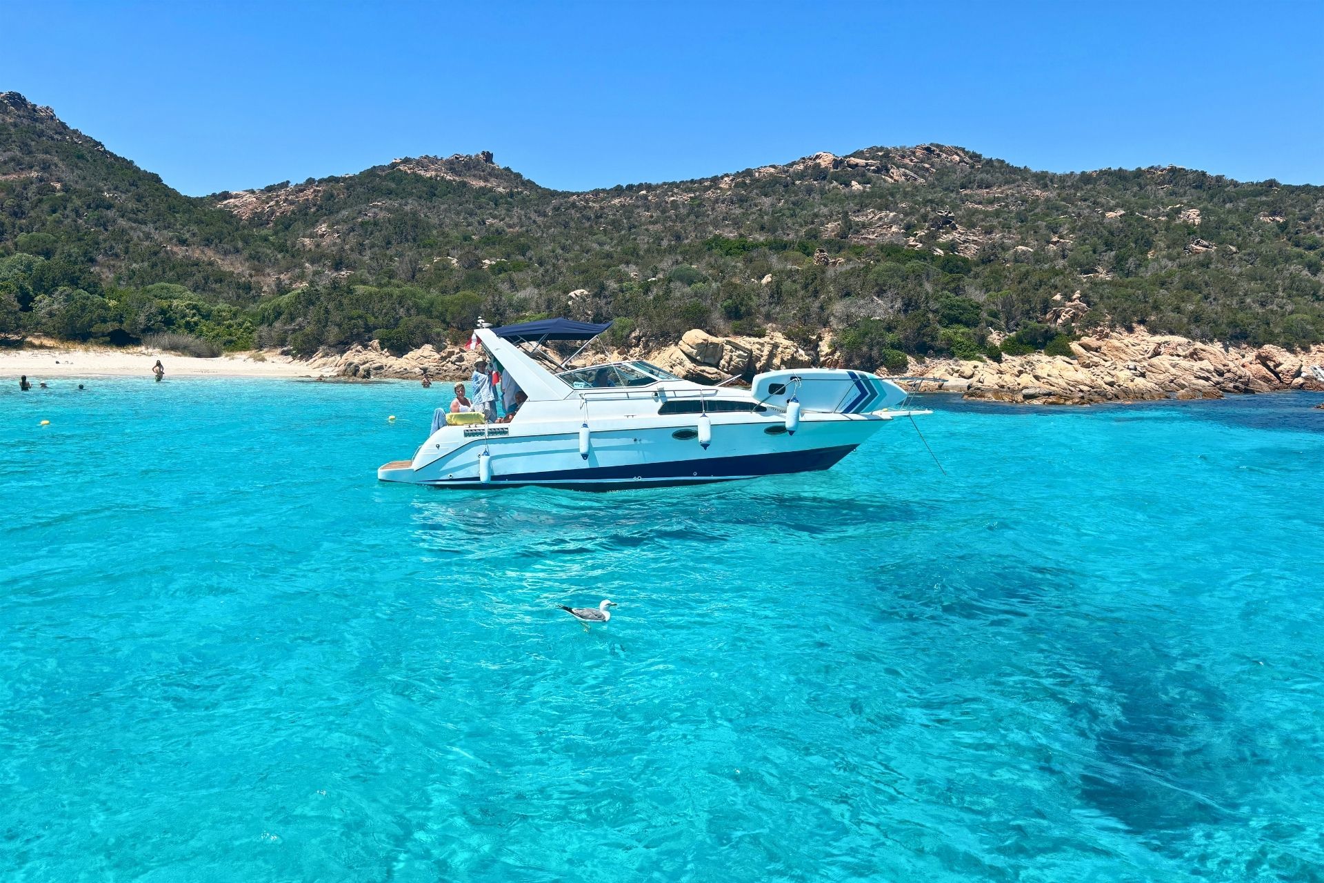 Image of a motorboat moored in crystal clear waters with people swimming and a surrounding natural landscape.