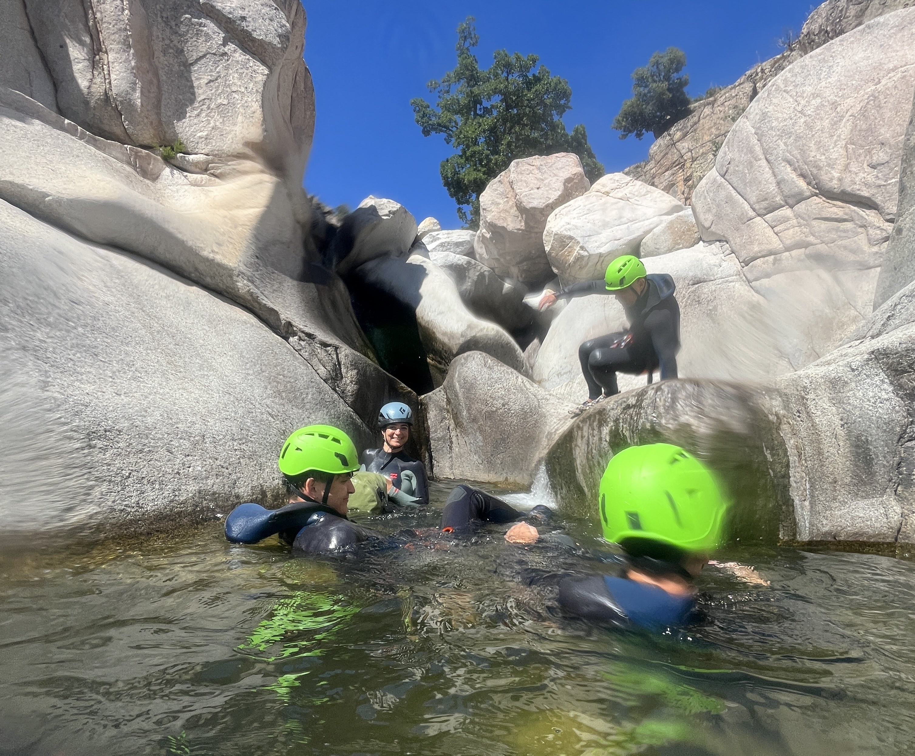 Persone in tuta e caschi immersi in un canyon con piscine naturali cristalline, circondate da rocce e vegetazione mediterranea